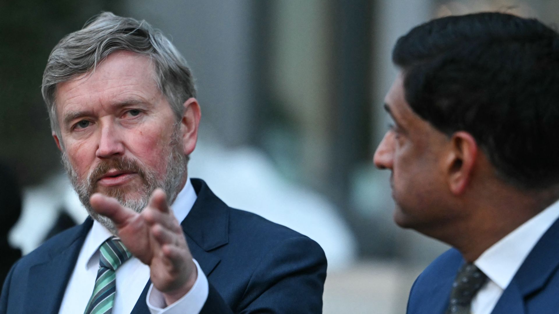 Two men in suits engaged in conversation outdoors; one man with gray hair and beard is gesturing with his hand, the other man has dark hair and is listening attentively.