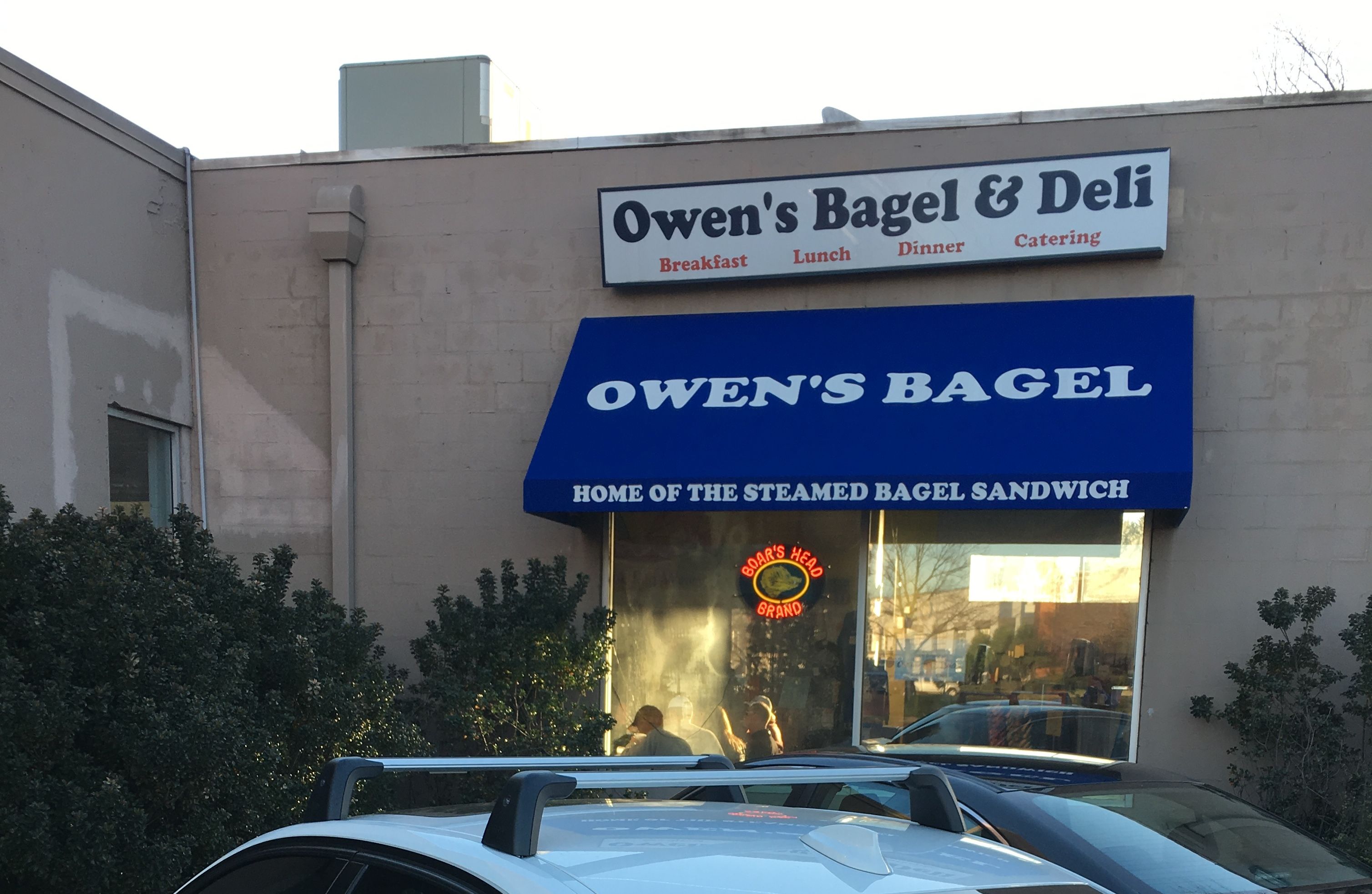 Exterior of Owen's Bagel & Deli with a blue awning reading "OWEN'S BAGEL" and "Home of the Steamed Bagel Sandwich." Sign above offers breakfast, lunch, dinner, catering. Cars and bushes in front.