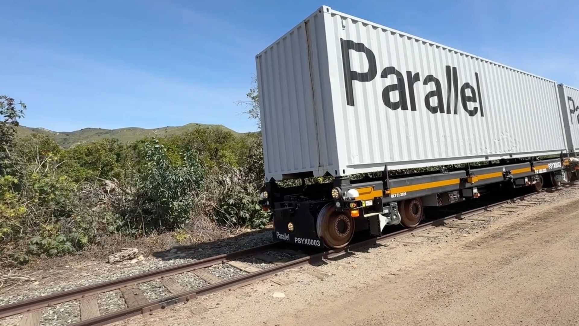 A white shipping container labeled Parallel rests on a rail chassis on railroad tracks, with scrubby bushes and distant green hills under a bright blue sky.