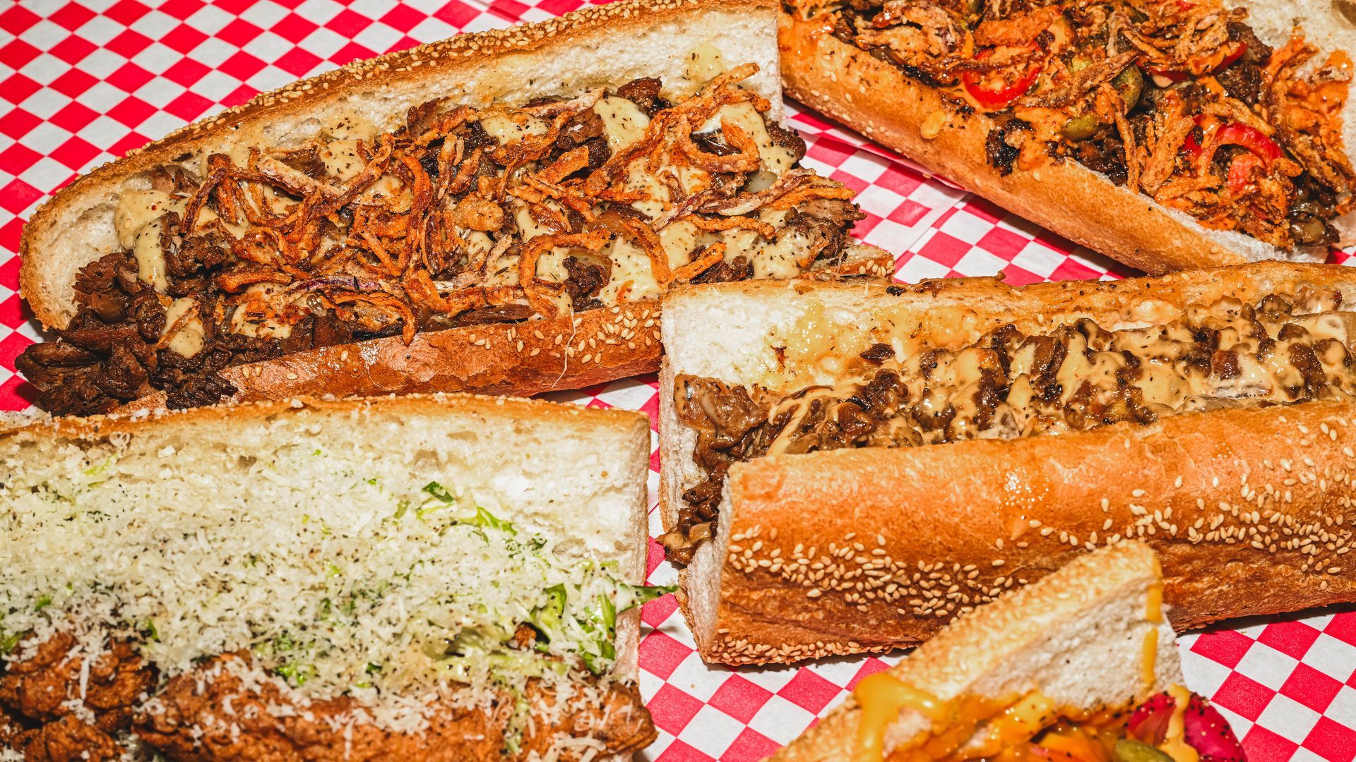 Assorted deli sandwiches on sesame seed hoagie rolls with shredded lettuce, melted cheese, fried onions, and grilled meat on red and white checkered paper.