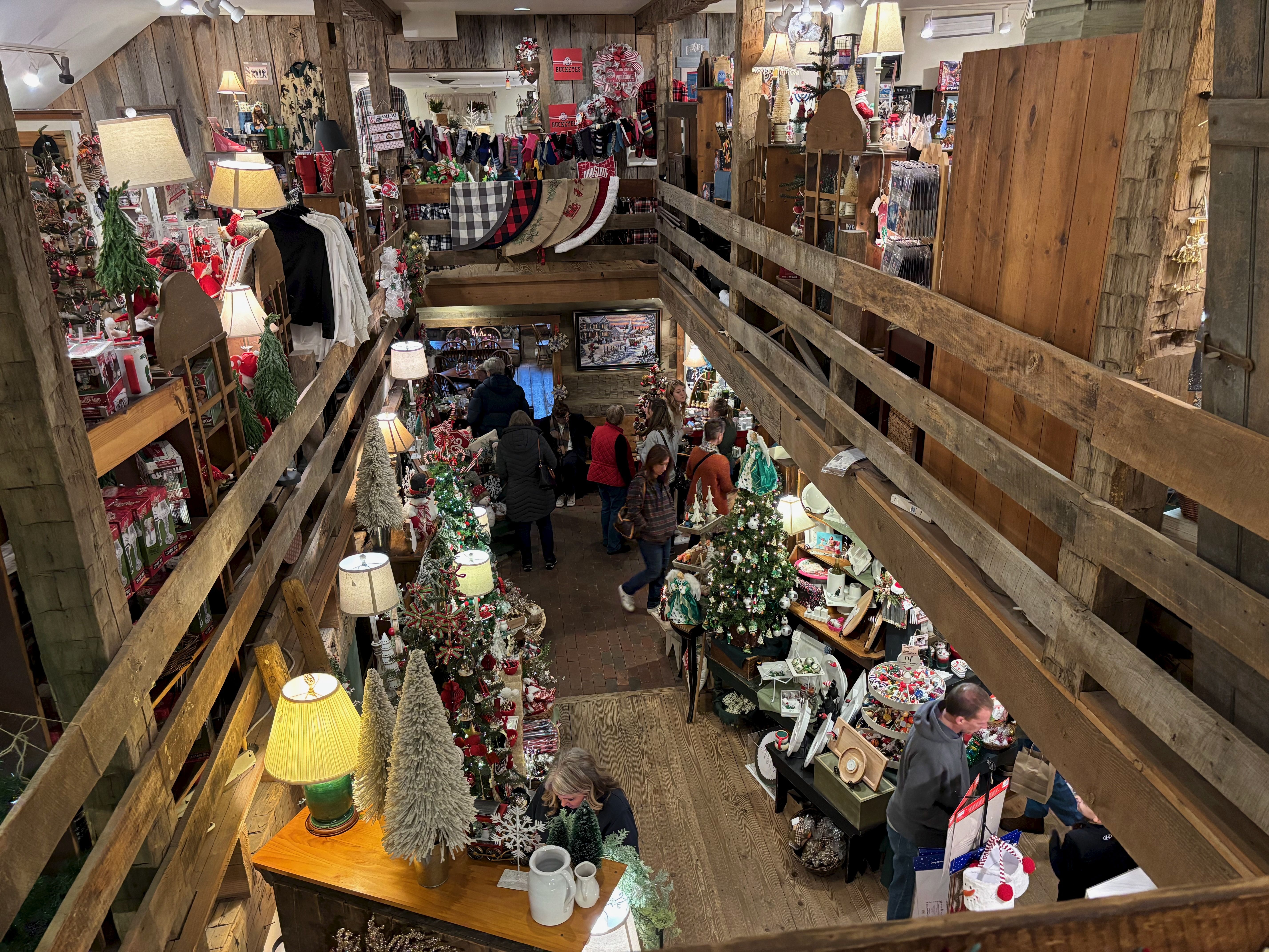 Cozy two-story gift shop decorated for Christmas with small decorated trees, lights, blankets, and holiday items, bustling with shoppers browsing and walking around.