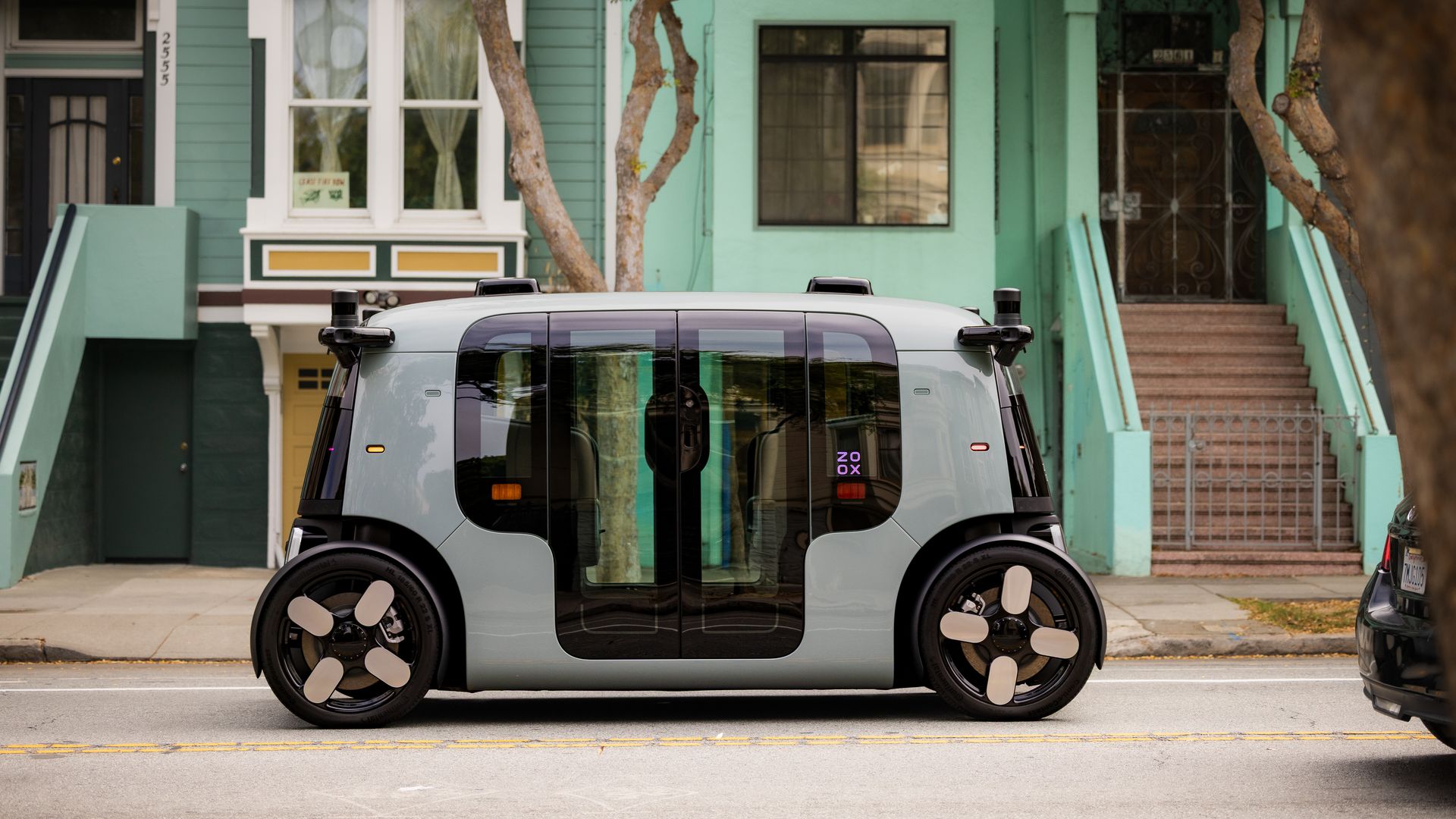 Side view of a compact, gray autonomous vehicle with black windows and unique four-spoke wheels parked on a city street in front of teal and green houses.