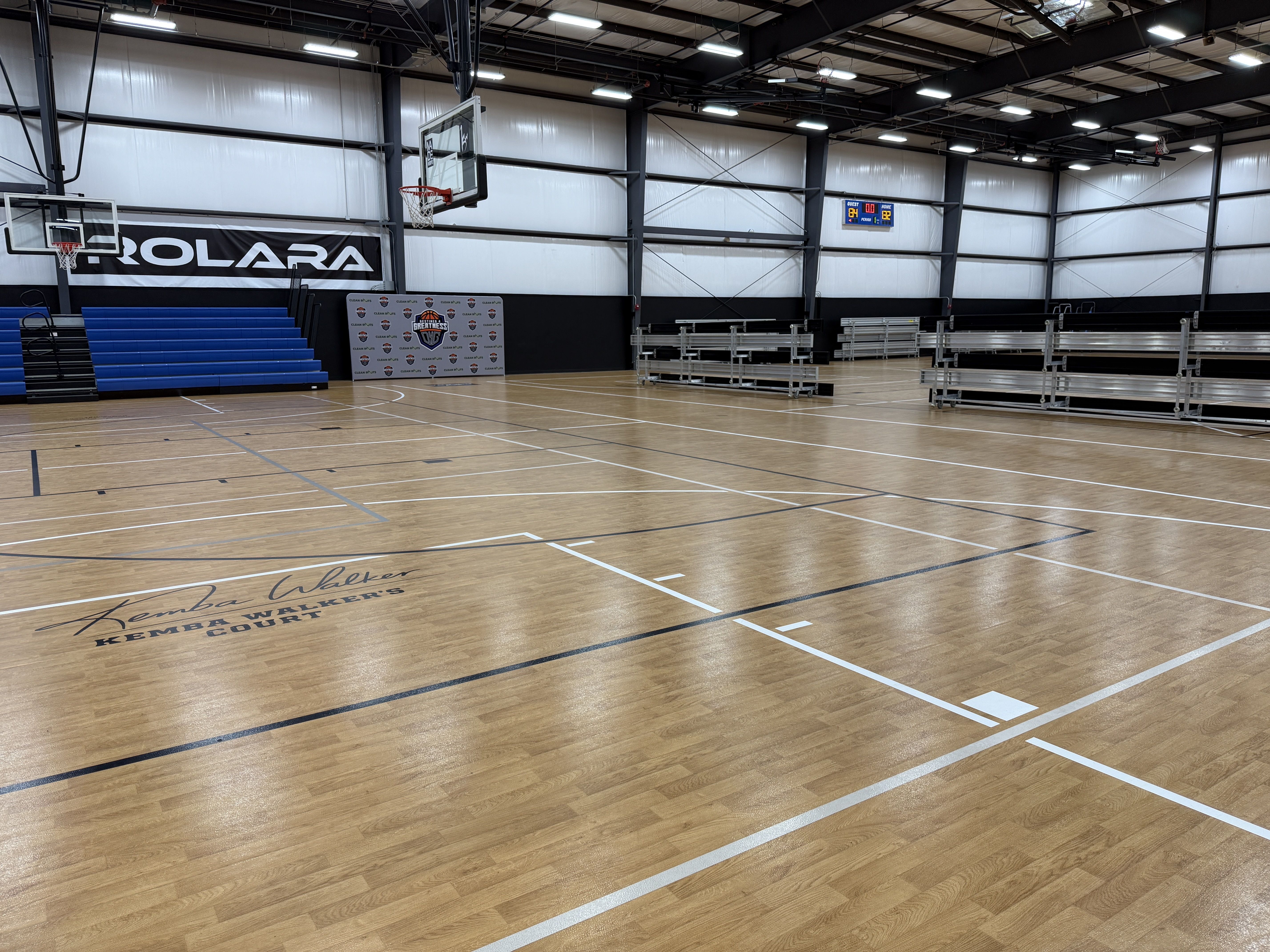 An empty indoor basketball court with a polished wood floor, blue bleachers on the left, two backboards and hoops, a large wall banner reading ROLARA, and a digital scoreboard in the background.