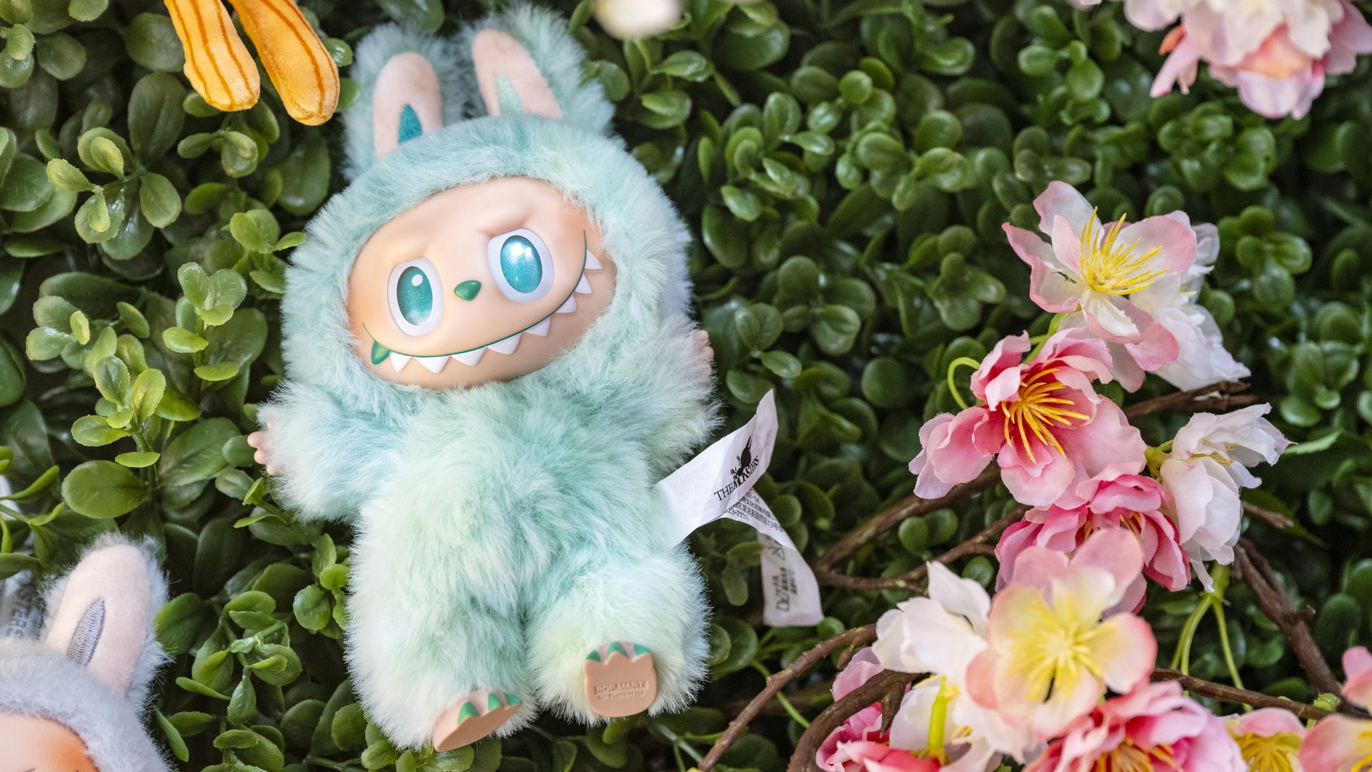 Light blue fluffy creature toy with big blue eyes and sharp teeth, surrounded by green leaves and pink flowers with yellow centers.
