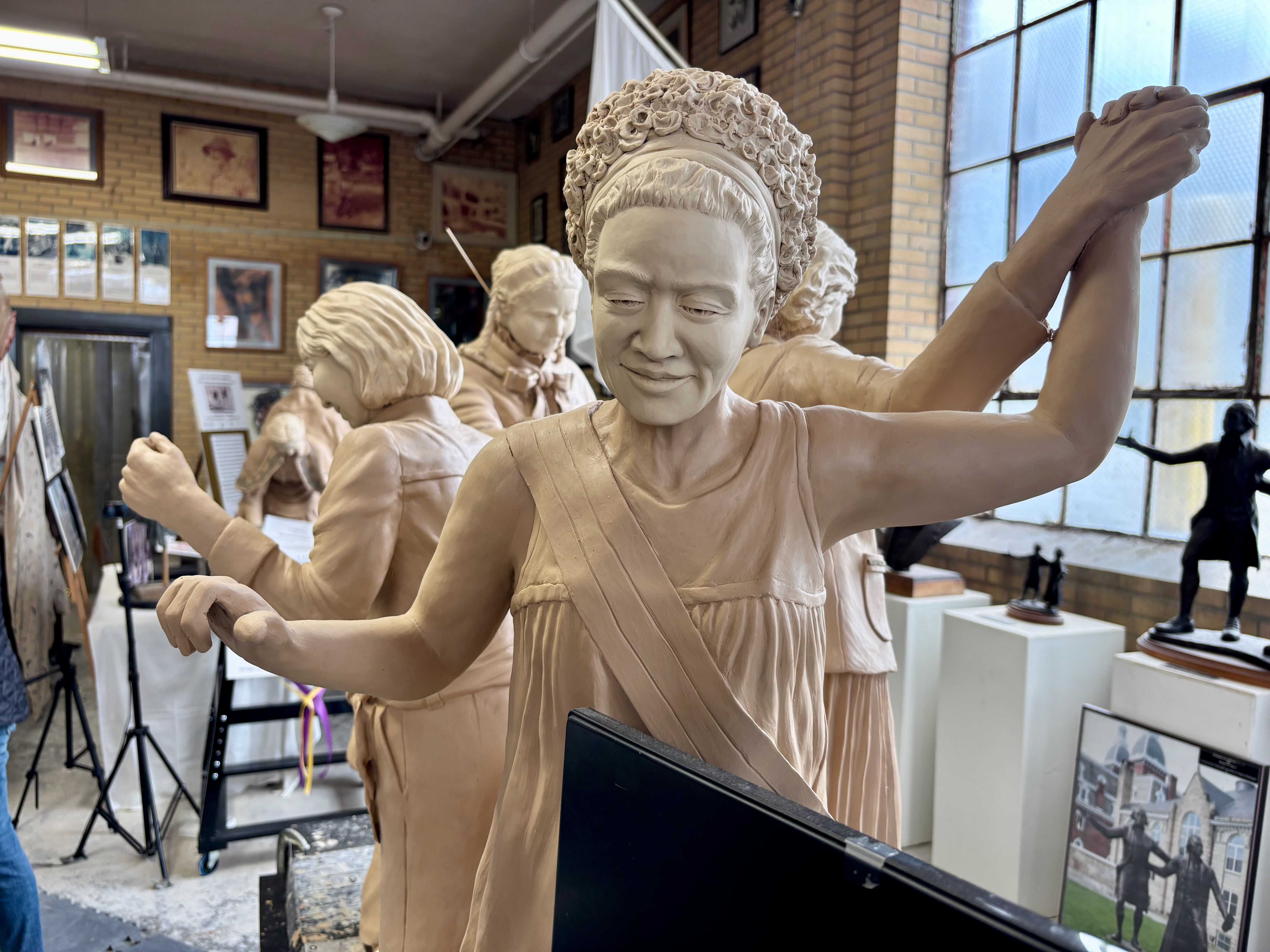 Indoor clay sculpture studio with pale figures; foreground shows a smiling woman raising her right arm, others in the background among framed art and brick walls.