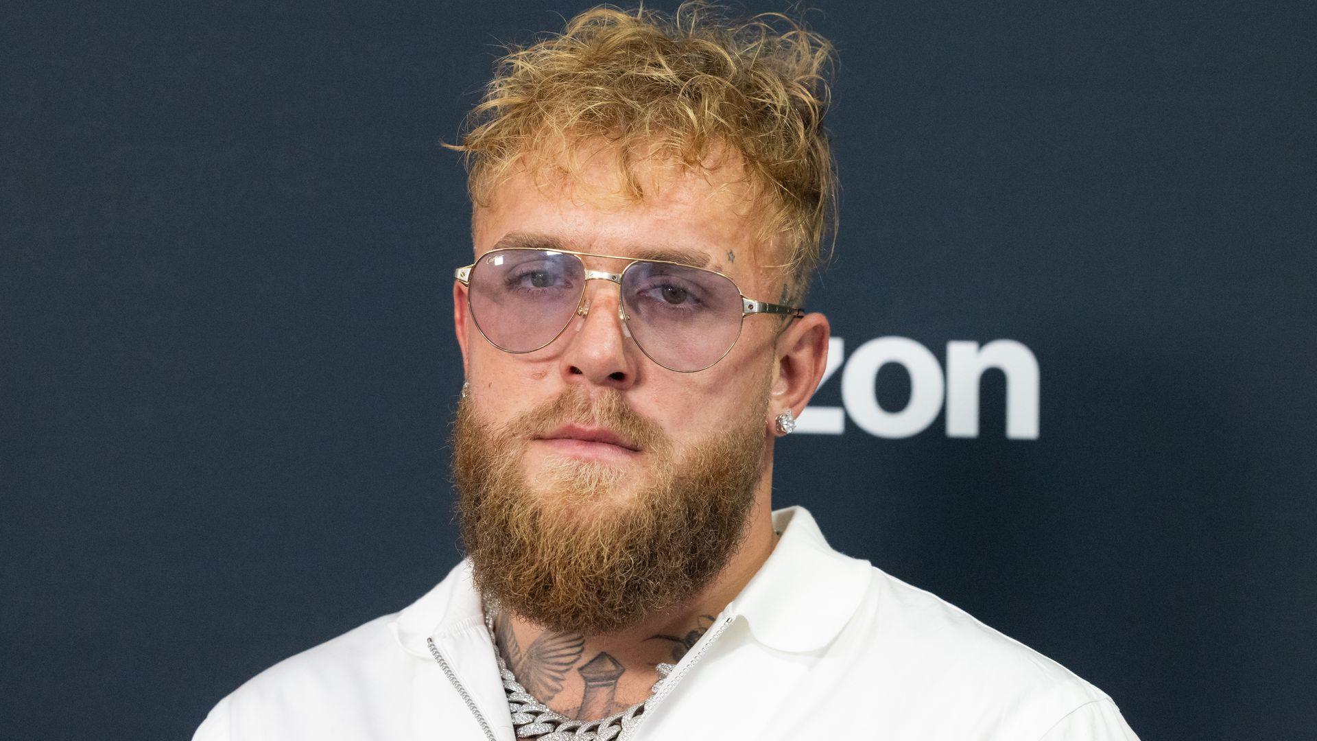 Portrait of a person with blond, spiky hair and a thick beard, wearing large aviator sunglasses, a chunky silver chain, and a white shirt. Neck tattoos visible; dark backdrop.