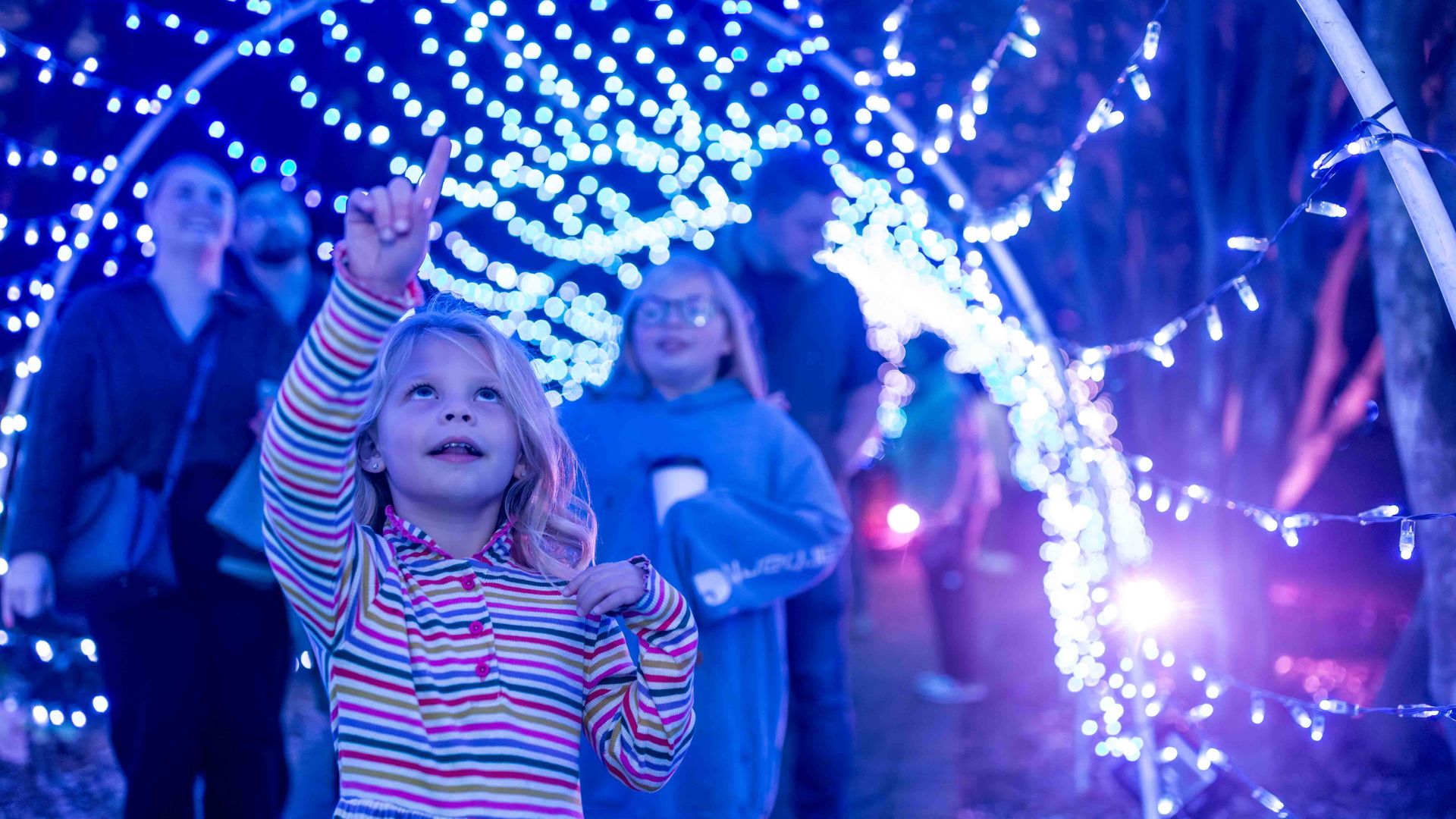 A young girl in a colorful striped dress points upward while standing under a tunnel of blue and white festive lights, with blurred people in the background.