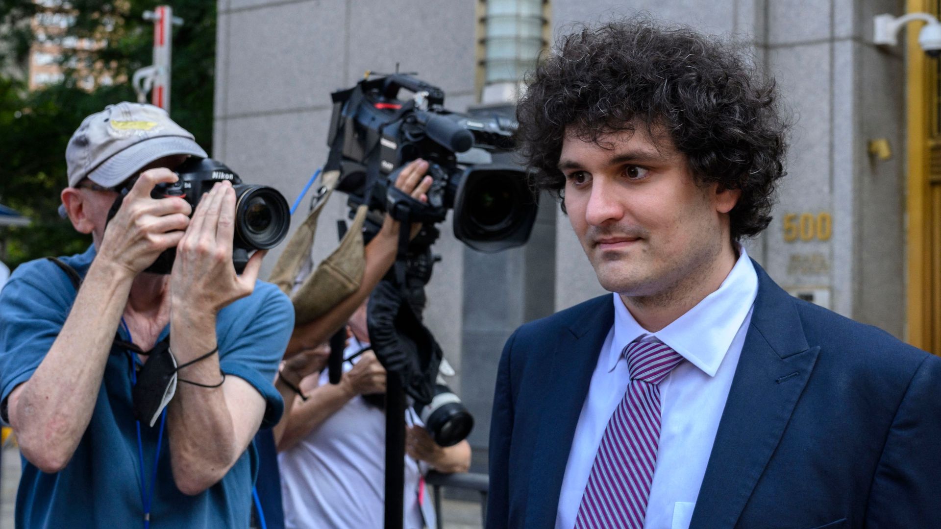 Sam Bankman-Fried, wearing a suit, exits a courthouse with cameramen taking shots behind him.