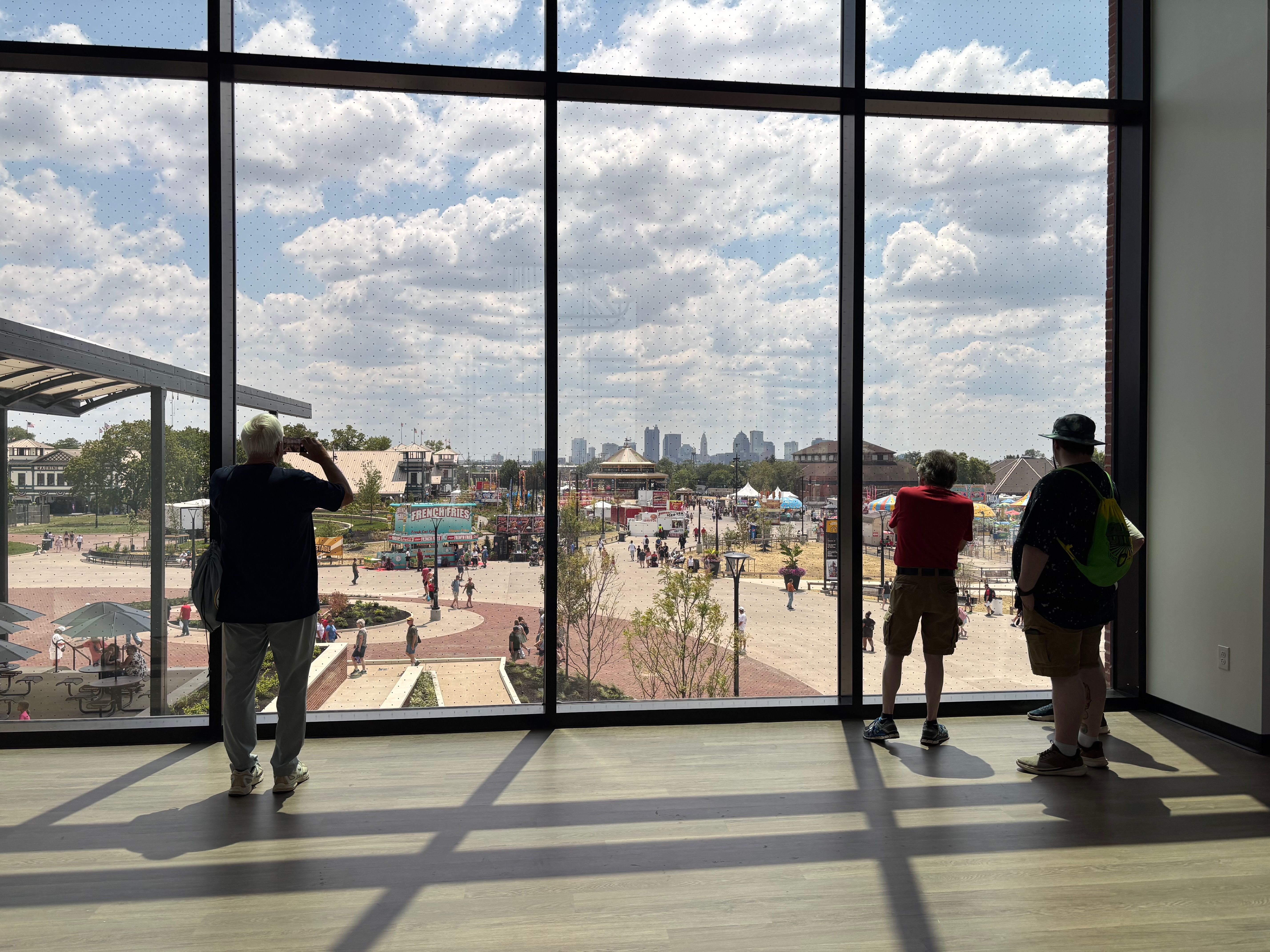 Inside a bright room with large grid windows and dotted glass, two people stand by the pane, overlooking a sunny outdoor fairground with rides, tents, and a distant city skyline.