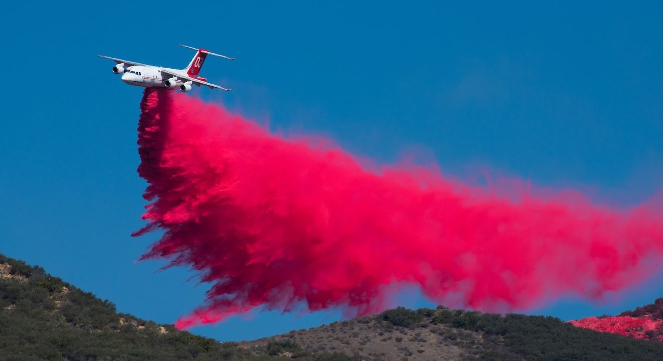 White passenger airplane emits a bright red smoke trail as it flies over low hills, creating a bold red plume across a clear blue sky.
