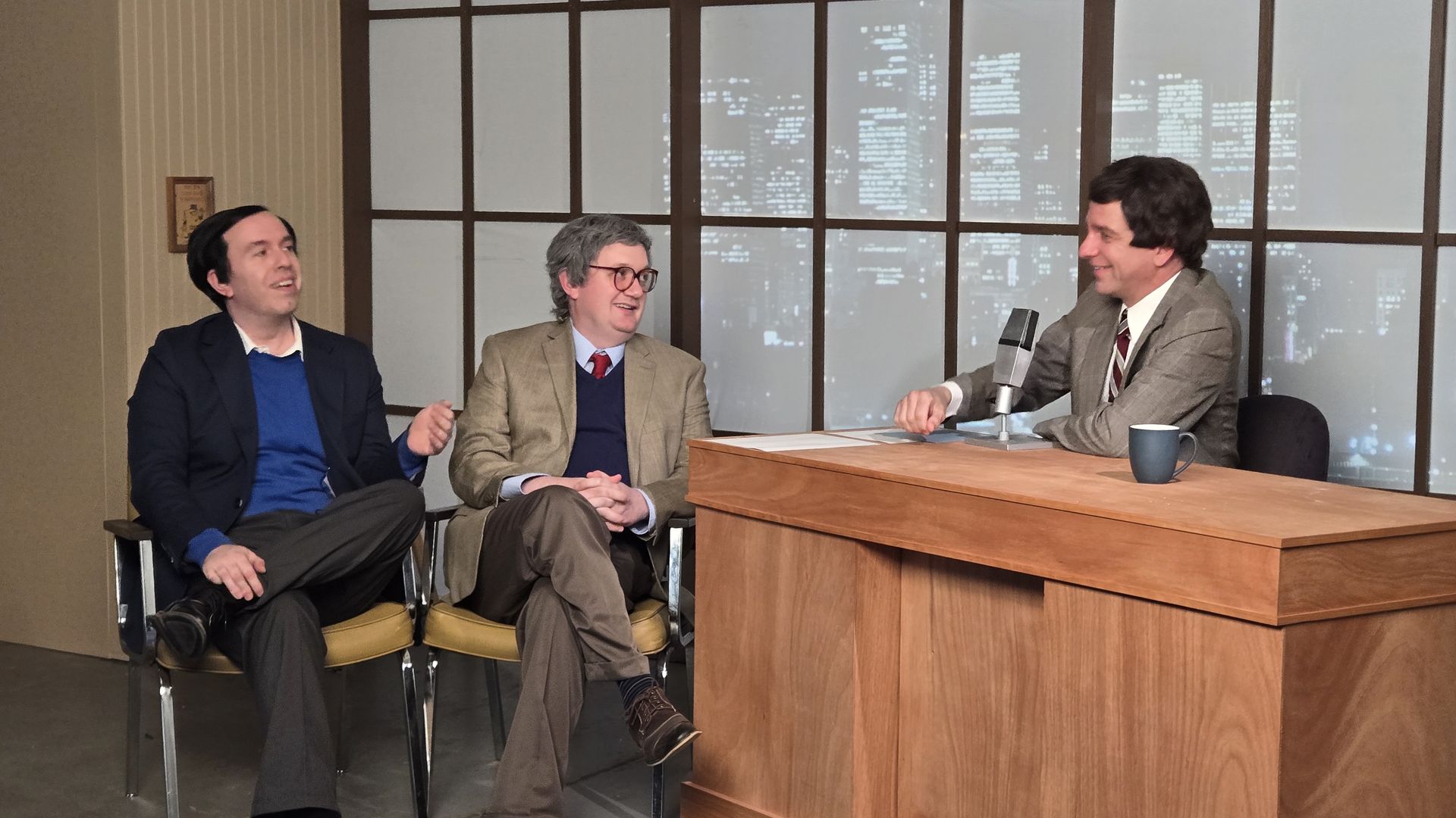 Three men in suits having a conversation on a talk show set with a wooden desk, microphone, blue mug, and city skyline backdrop at night.