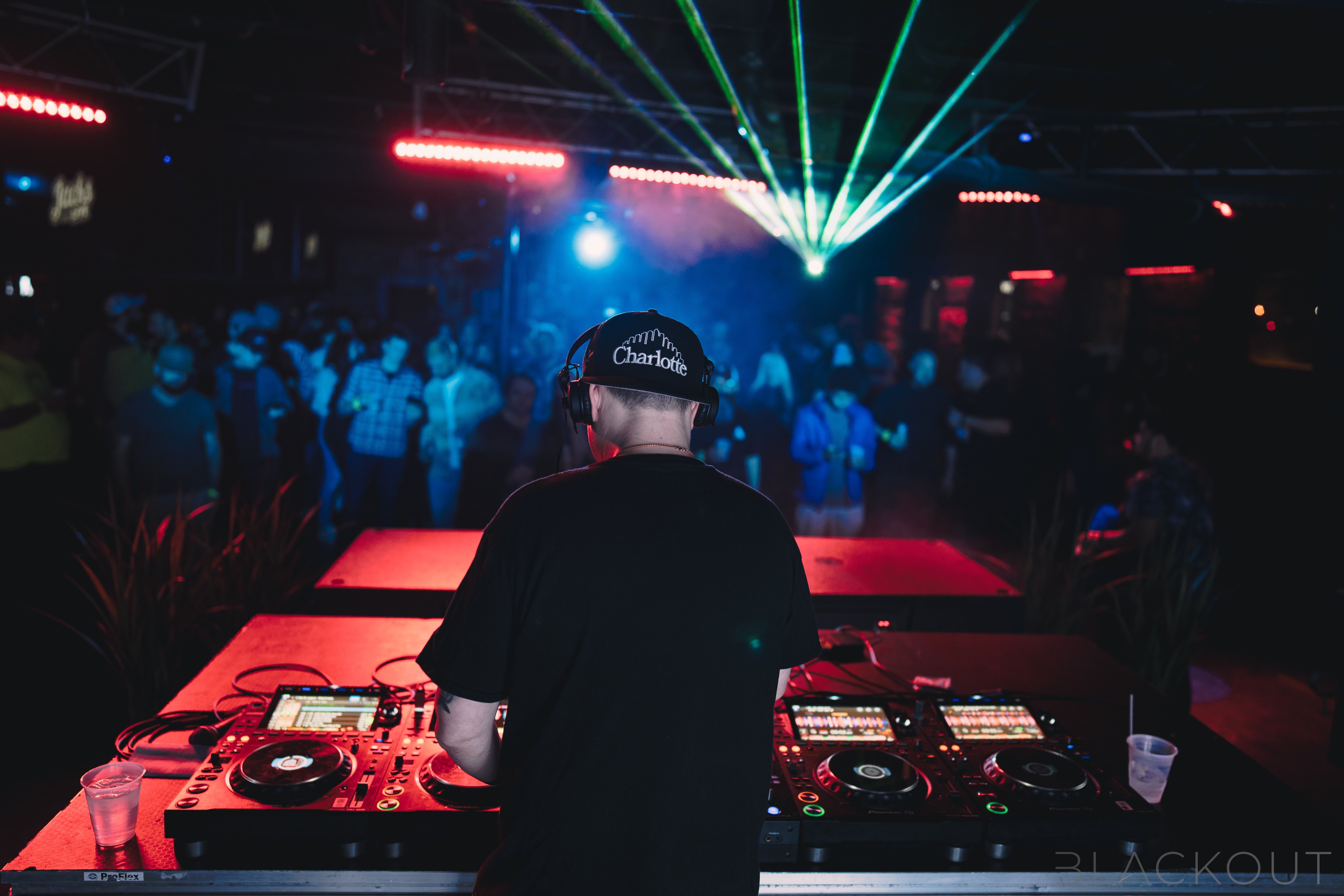 DJ wearing a black "Charlotte" cap and headphones, performing at a club with red lights, green lasers, and a crowd in the background.