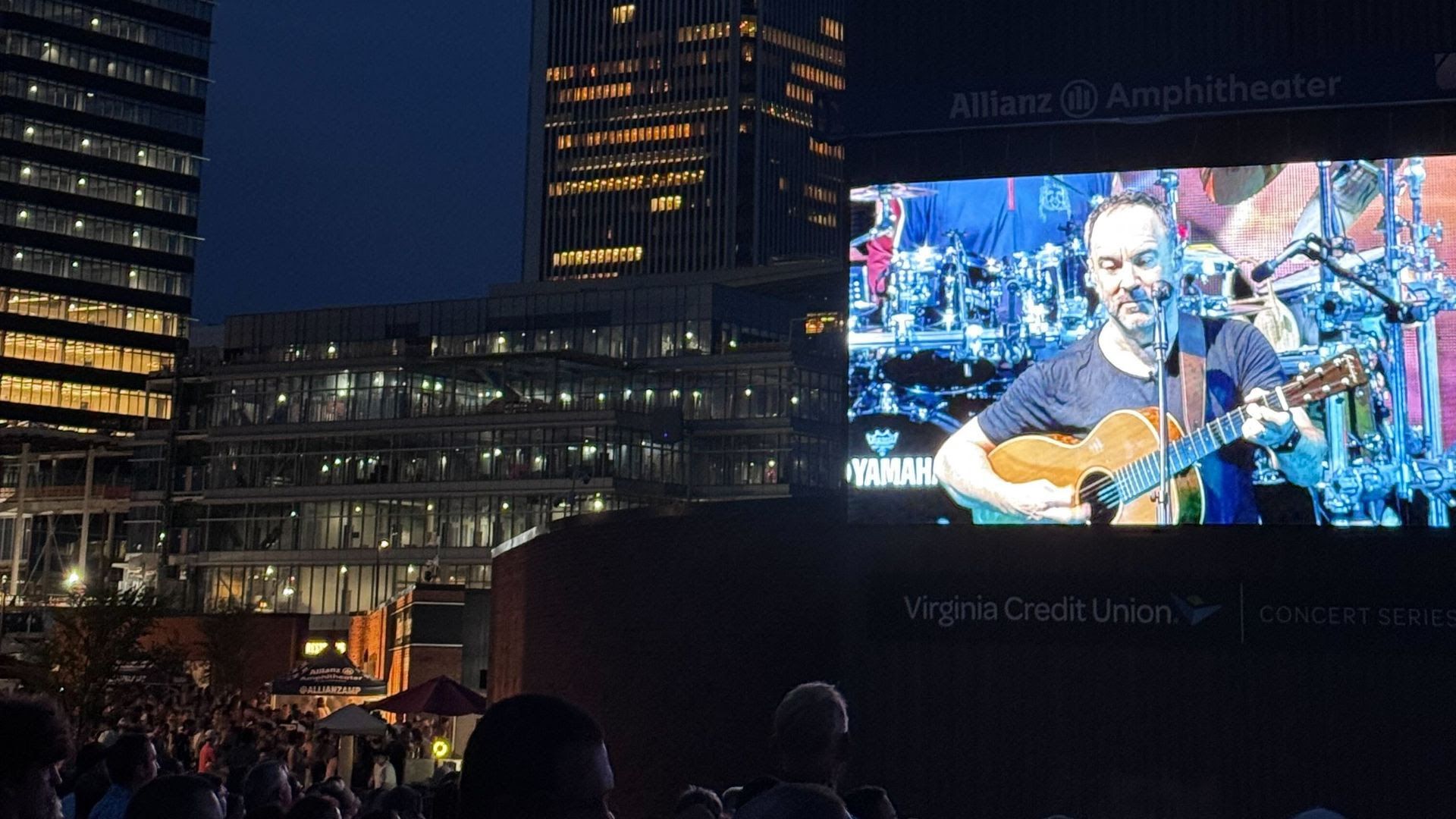 Nighttime outdoor concert at Allianz Amphitheater showing a large screen with a musician playing guitar, surrounded by buildings with lit windows and audience silhouettes.