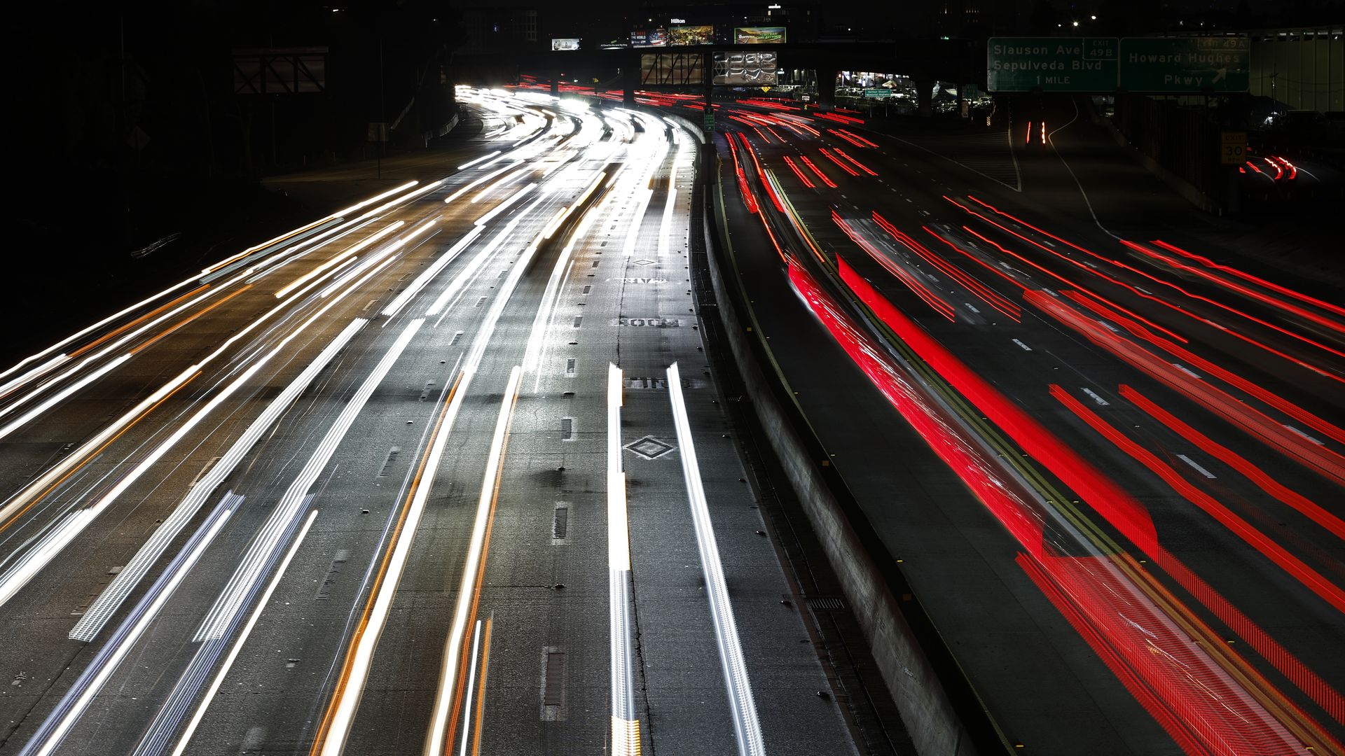 Nighttime highway with white light trails on the left and red taillights on the right, concrete median barriers, and green overhead signs; distant city lights glow.