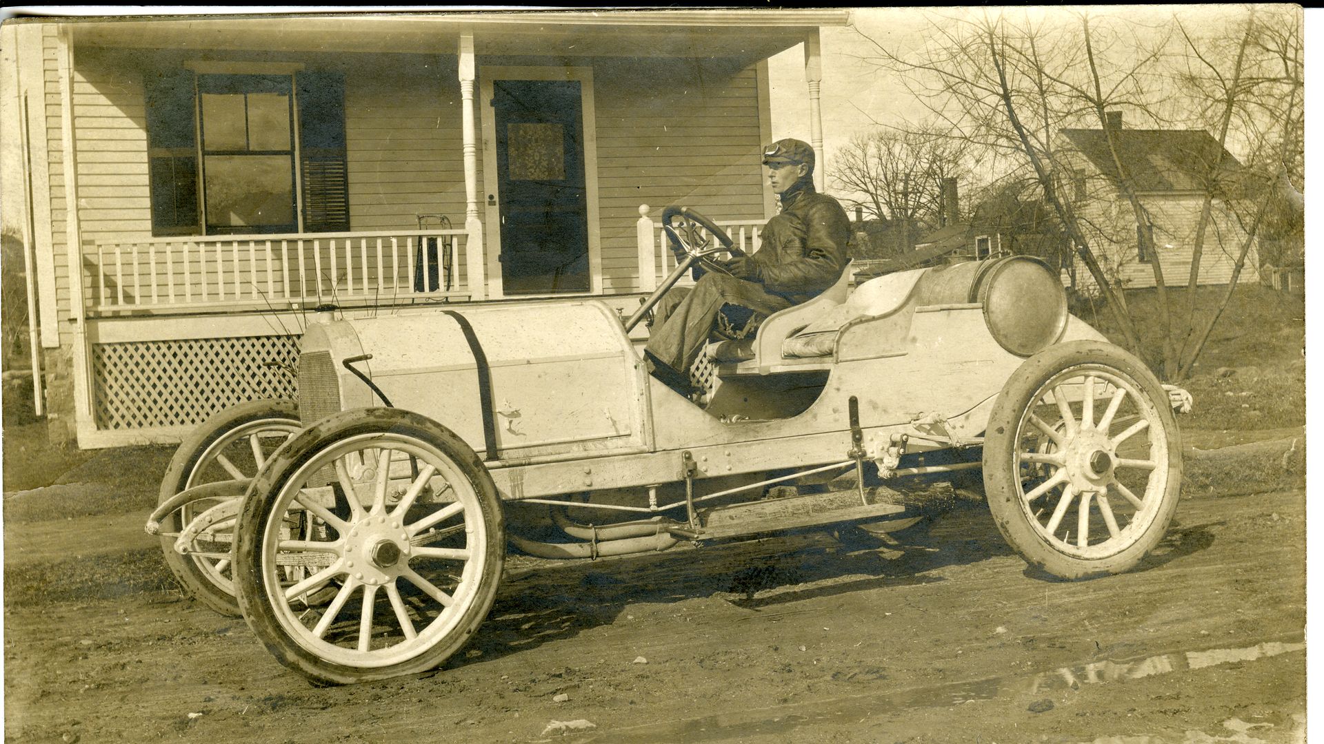 Sepia-toned vintage open-top car with large wooden-spoked wheels parked in front of a wooden house porch; a man in a cap sits at the steering wheel, with leafless trees in the background.