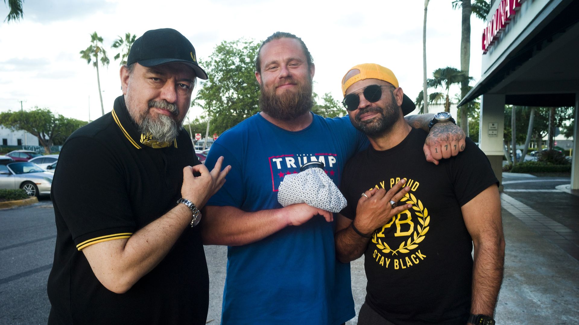 Three men pose in a parking lot, smiling. Left in black polo and cap, middle in blue shirt with a Trump logo and beard, right in Proud Boy black tee with a yellow cap and sunglasses. Palm trees and a building behind.