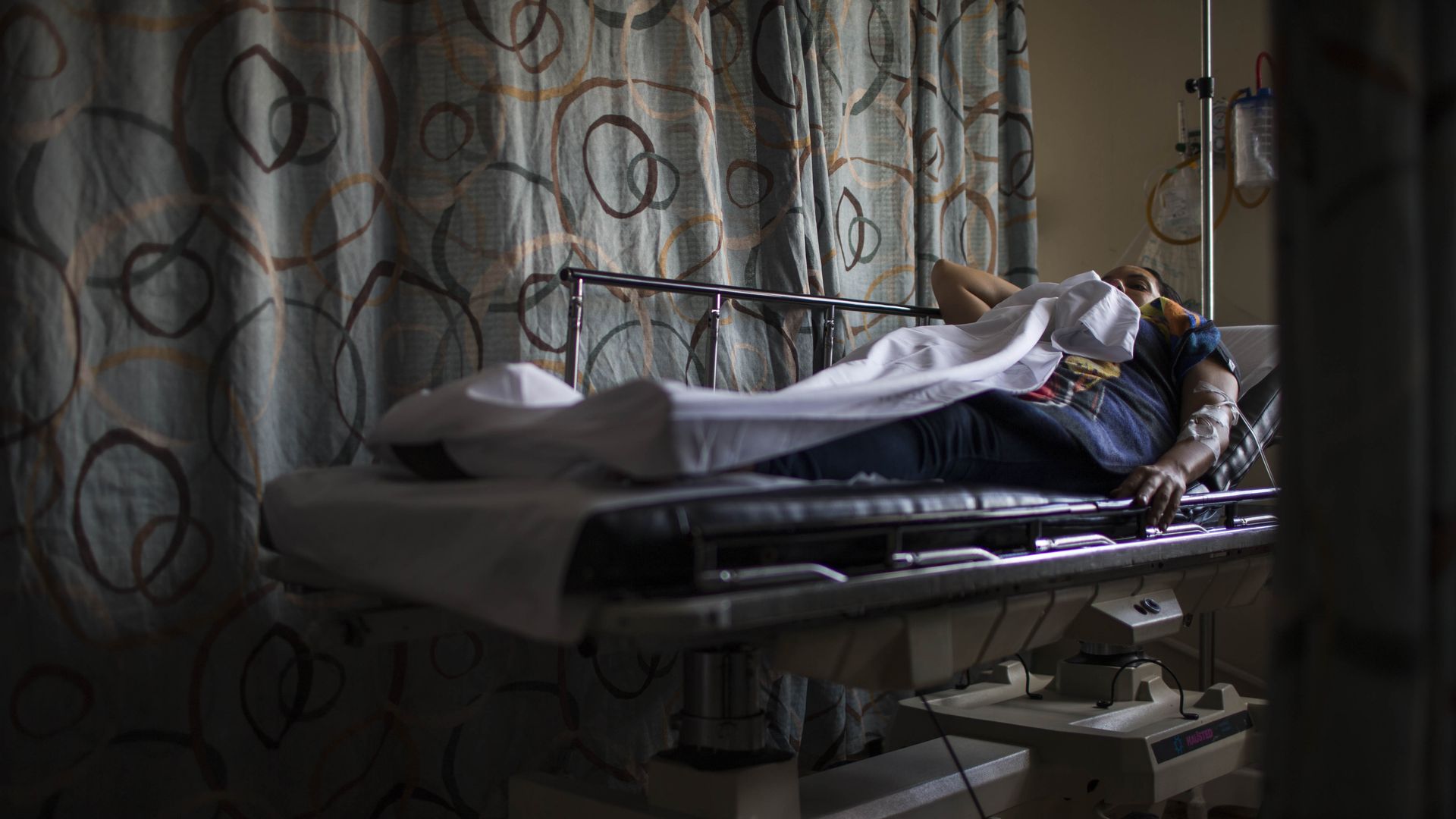 A person lays on a hospital bed in a dark emergency room.