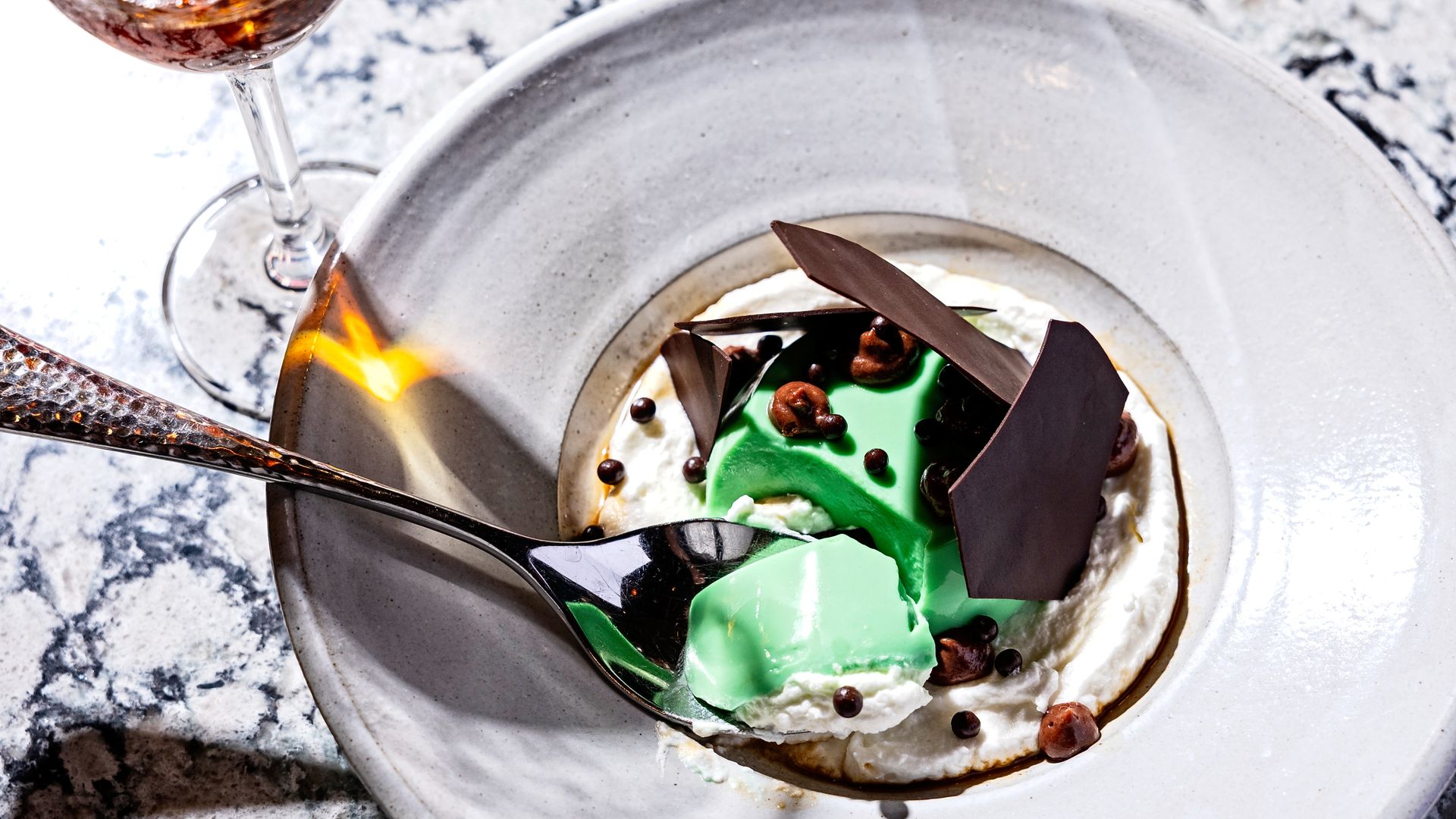 White plate with green mousse dessert topped with dark chocolate shards and small chocolate spheres, sitting on whipped cream, with a silver spoon and a glass of amber liquid on a marble surface.