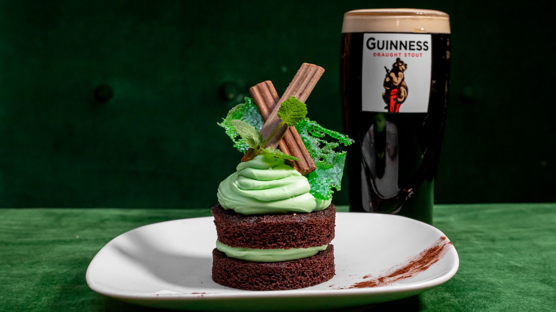 A dessert with two chocolate cake layers filled and topped with mint-green frosting, garnished with chocolate sticks, mint leaves, and green sugar lace, on a white plate beside a pint of Guinness.