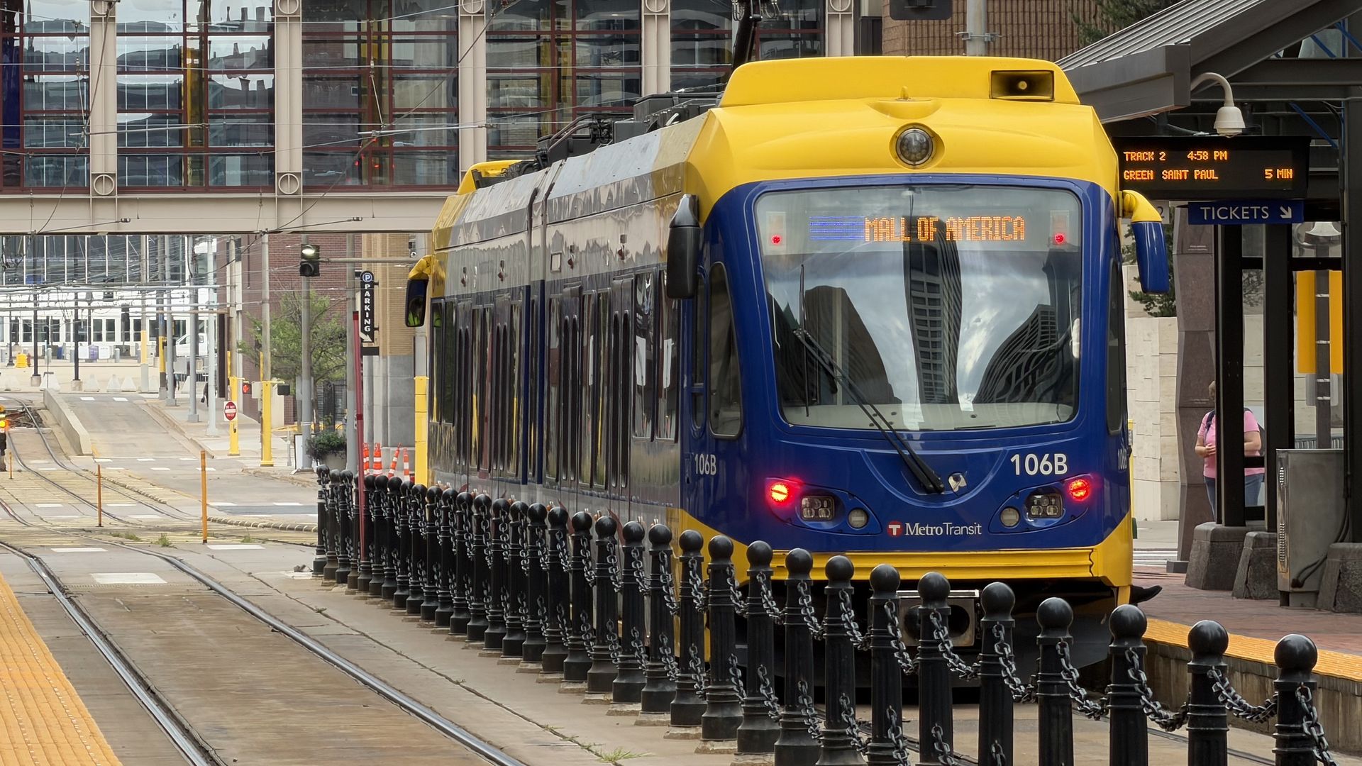 Blue and yellow tram at a modern city station with overhead wires, glass office towers in the background, and rails; the platform shows yellow tactile paving; a pedestrian is visible on the right.
