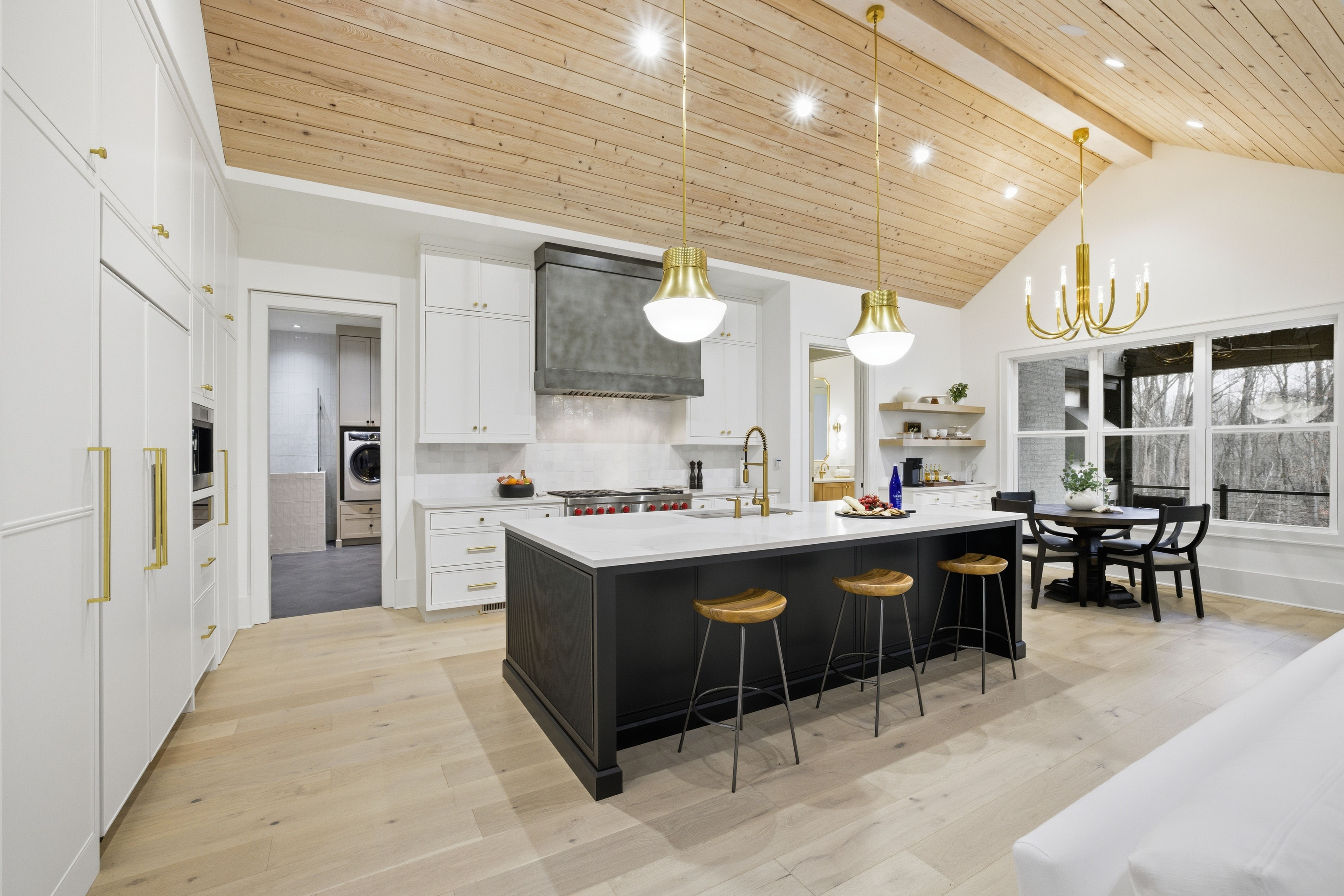 Bright modern kitchen with black island, wooden stools, white cabinets, brass fixtures, light wood floors, wood-paneled ceiling, and dining area with black chairs and large windows.