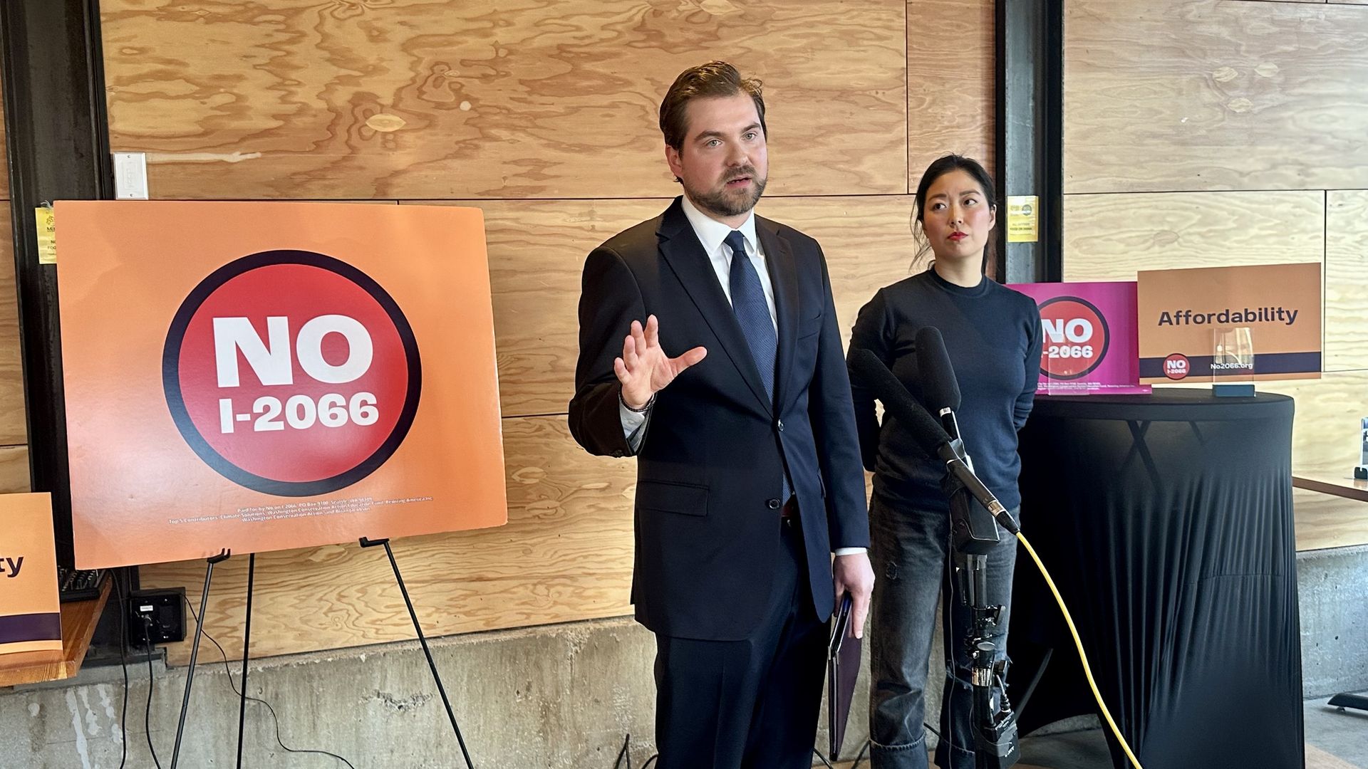 A man in a suit stands behind a microphone speaking with his hands moving as he speaks, with a "No I-2066" sign to his right and a woman standing behind him to the side.
