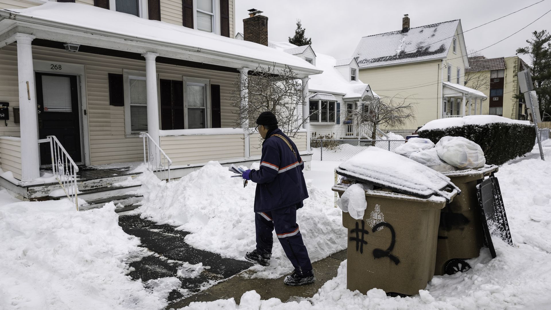 Snow-covered residential street with a mail carrier in blue uniform delivering mail, two trash bins marked #2, and beige houses with snow on roofs and sidewalks.