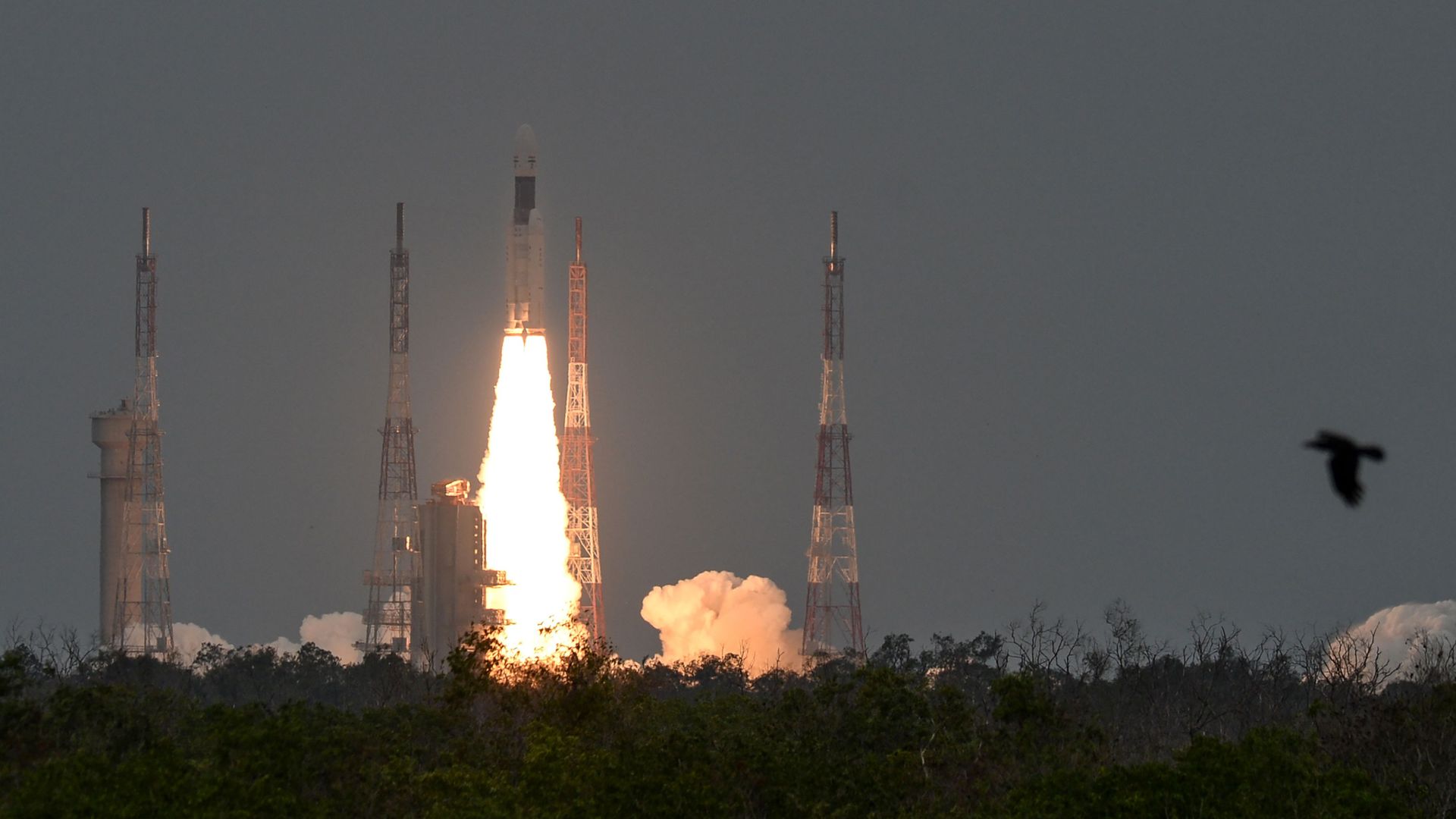 In this image, a rocket launches between four radio towers, with forest in the foreground.