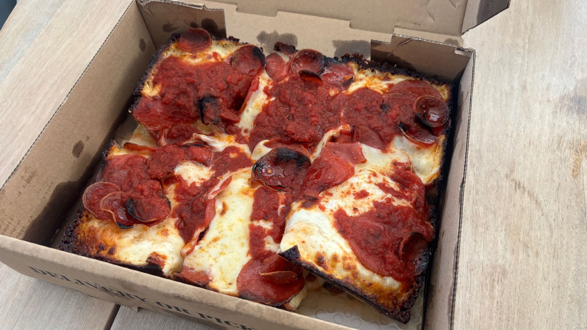Pepperoni pizza in an open cardboard box on a wooden table; cheese melted, pepperoni slices scattered, with dark burnt edges along the crust.
