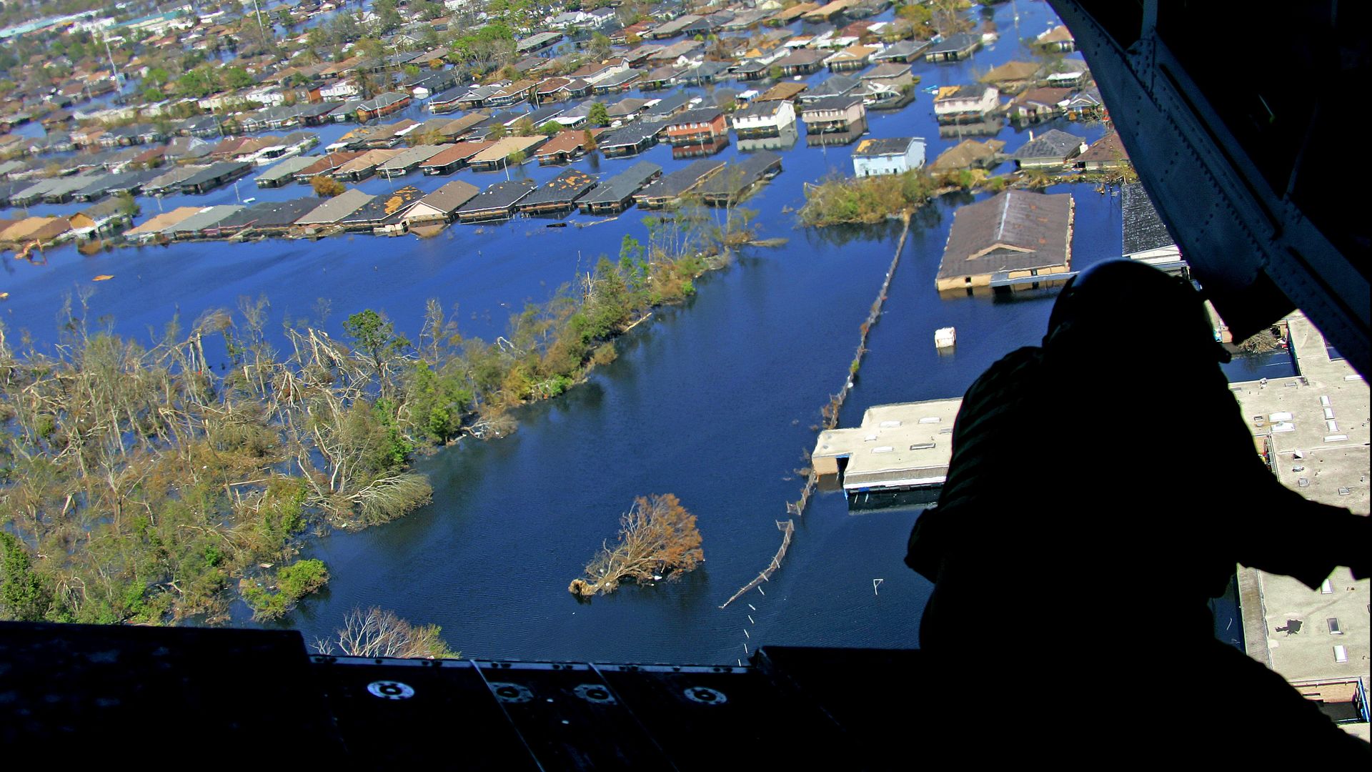 Aerial view of a flooded residential area with submerged houses and trees, seen from a dark aircraft interior with a silhouetted person leaning out.