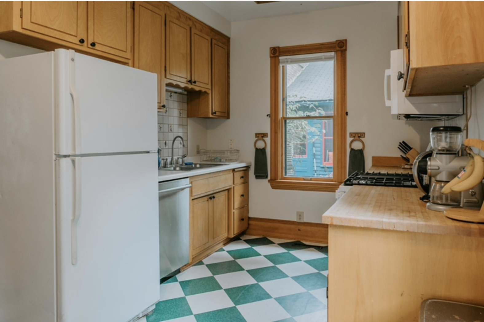 Small kitchen with wooden cabinets, white fridge, dishwasher, gas stove, checkerboard green and white floor, window with brown trim, coffee maker, and bananas on counter