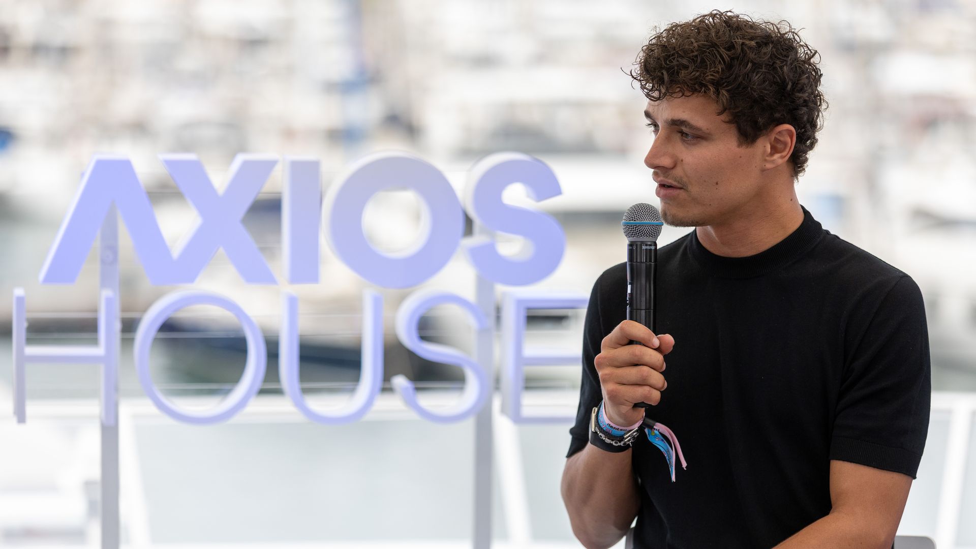 Side-profile of a young man with curly dark hair in a black shirt, holding a microphone and speaking; blurred harbor backdrop with large lavender letters behind him.