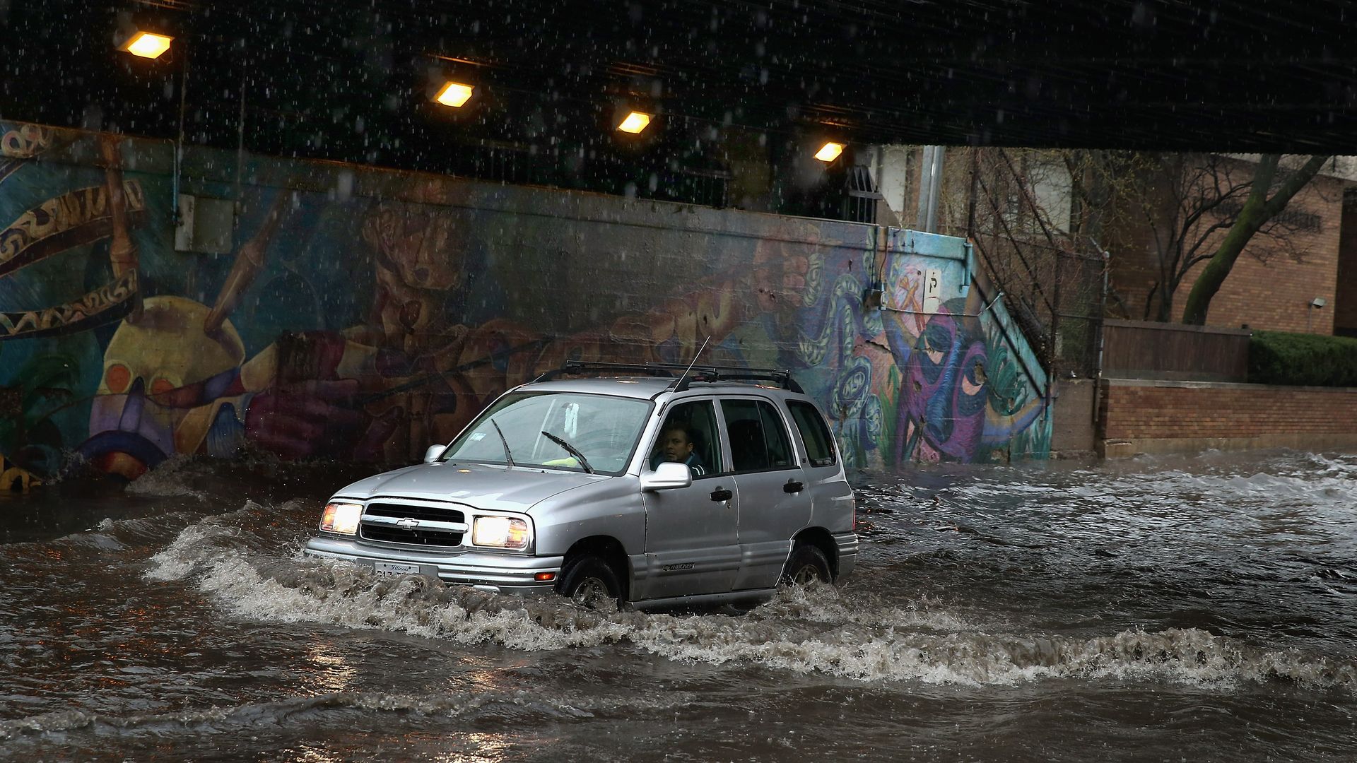 Silver SUV driving through floodwater under a graffiti-covered underpass in heavy rain; water splashes around its wheels while orange lights glow overhead.