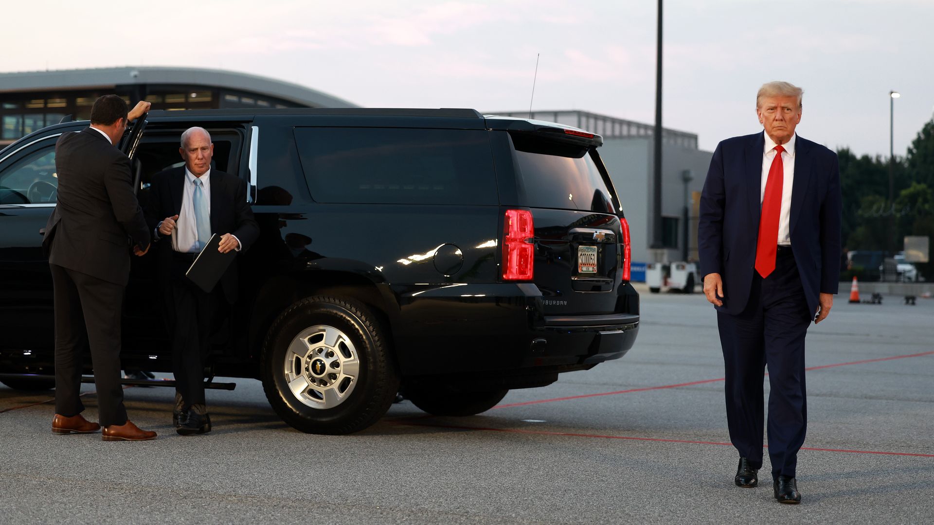 Trump on the right side of the photo in a suit and red tie walking away from a black car. To the left, his attorney in a suit and blue tie gets out of the car. 