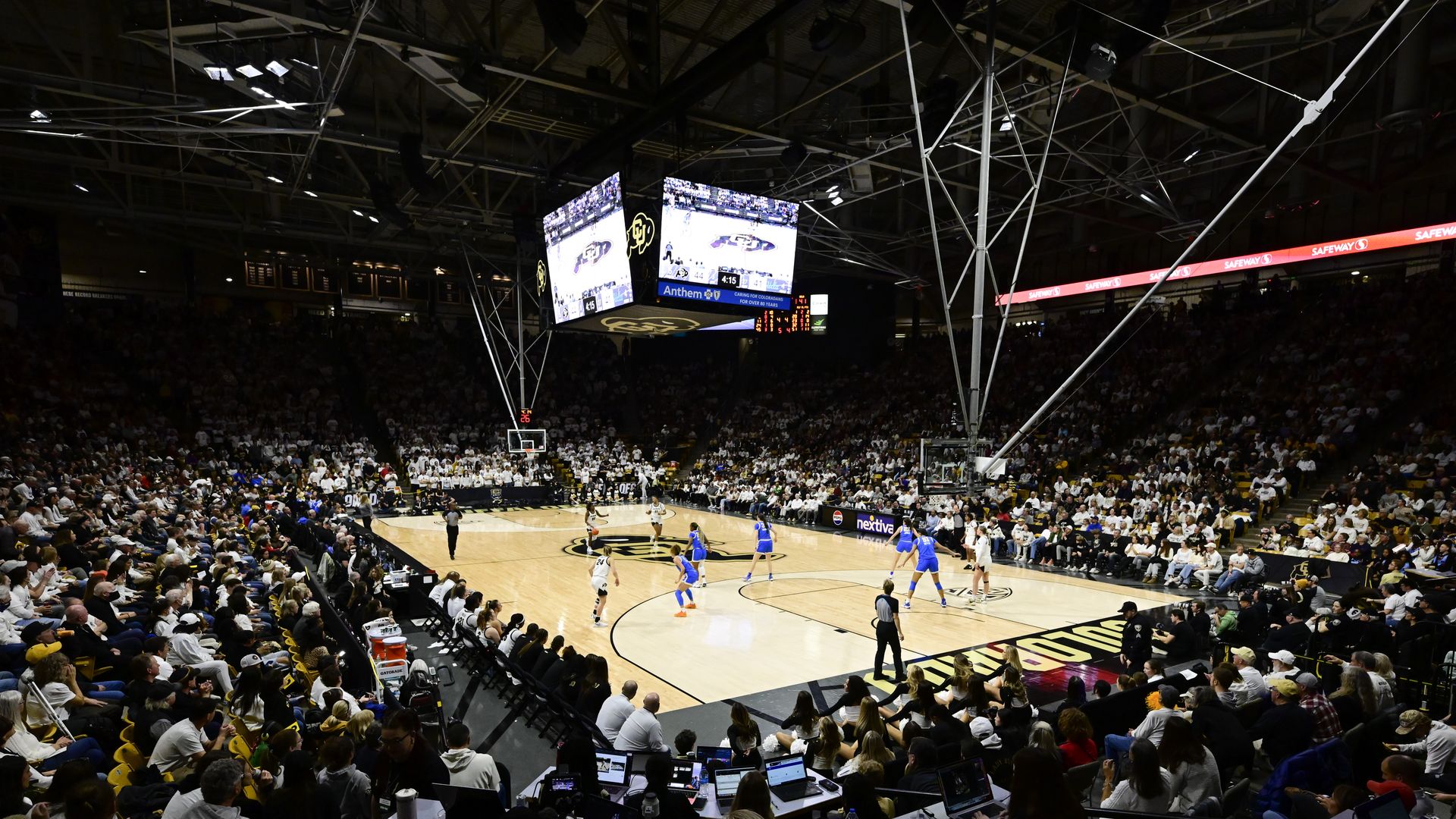 Crowded basketball arena with players on court, large central scoreboard and hanging screens, bright lighting, and white-clad fans filling the stands.
