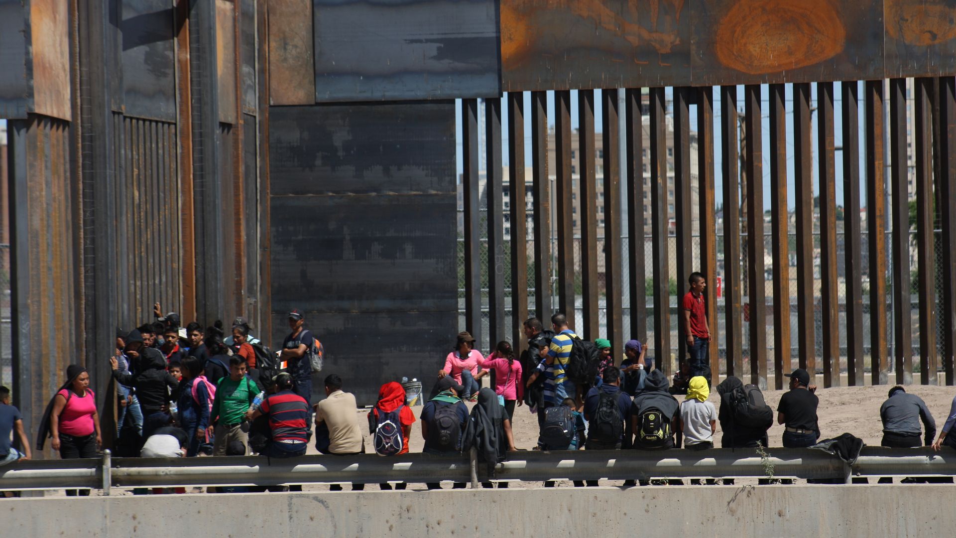 A crowd of migrants standing and sitting outside of a large border fence on the southern border. 