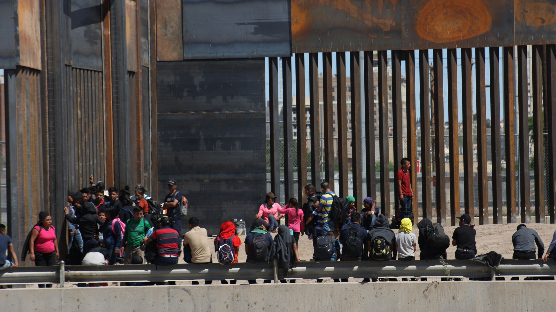 A crowd of migrants standing and sitting outside of a large border fence on the southern border.