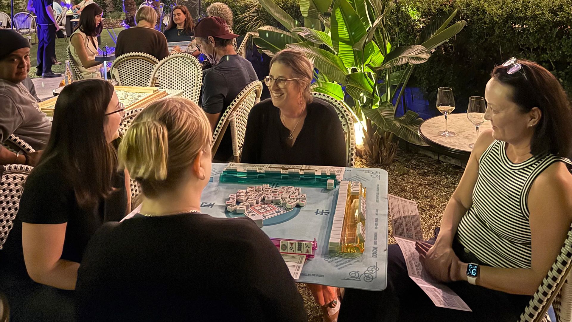 Night outdoor gathering with people sitting at tables playing Mahjong under string lights, surrounded by palm trees and a large house lit with blue lights in the background.