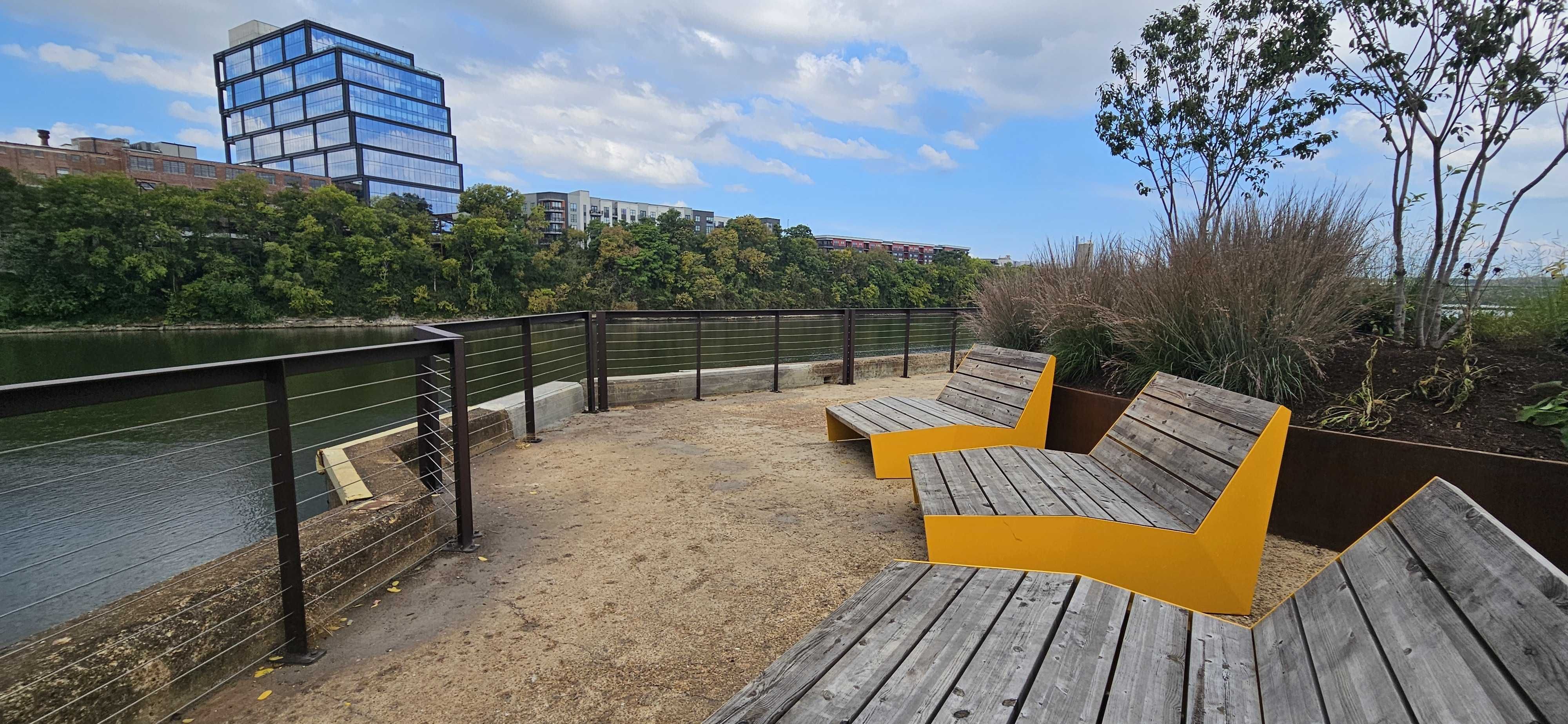 Riverfront park with wooden benches that have bright yellow angular bases, a metal railing along the water, trees in the background, and a modern glass building under a blue sky.