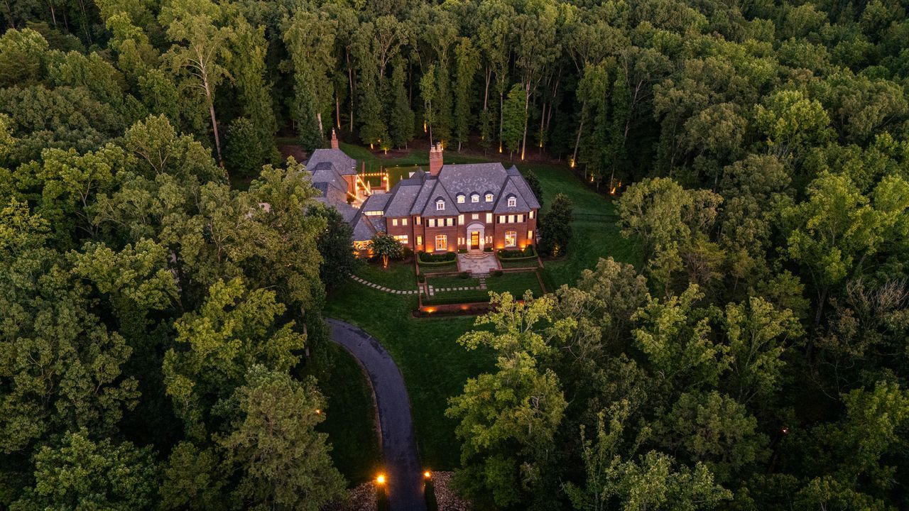A large, illuminated brick house with a slate roof sits surrounded by dense green forest, visible from a curved driveway at dusk.