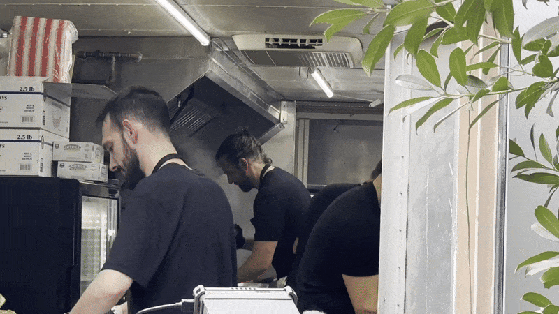 Bradley Cooper uses a spatula at a crowded food truck to chop up beef. His back is turned to the camera and he's on the far side of the truck, viewed through the back door.