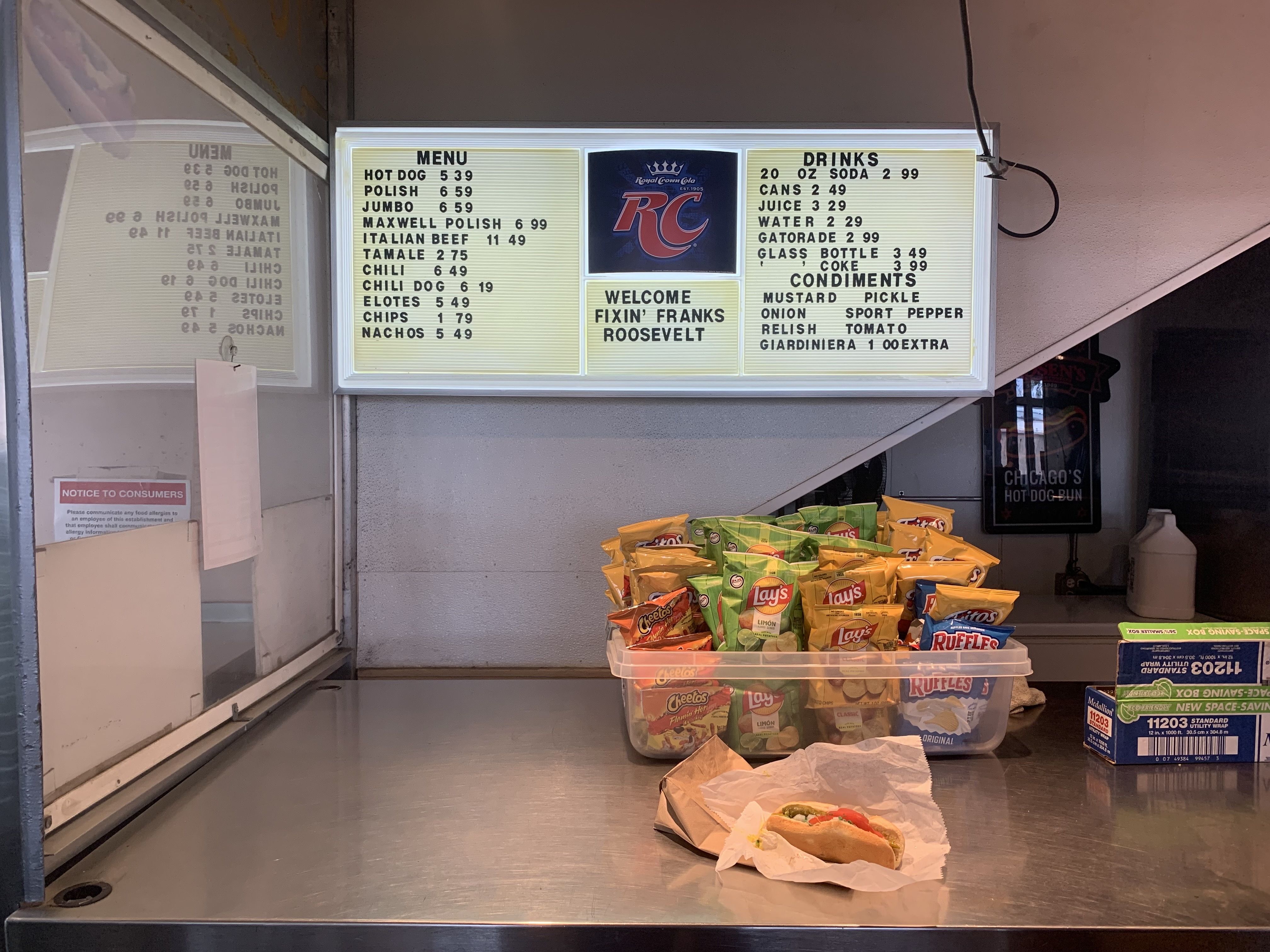Illuminated retro menu board at a snack counter; left column lists hot dogs and sides, right column drinks; a blue RC logo center panel; chips and a wrapped hot dog sit on the counter below.