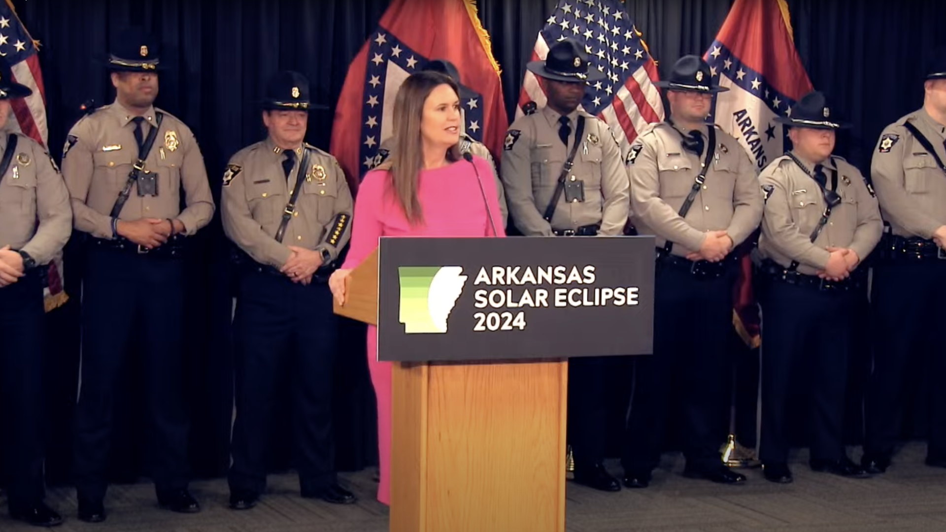 A photo of Arkansas Gov. Sarah Huckabee Sanders standing at a podium with a sign reading "Arkansas Solar Eclipse 2024" and flanked by state troopers.