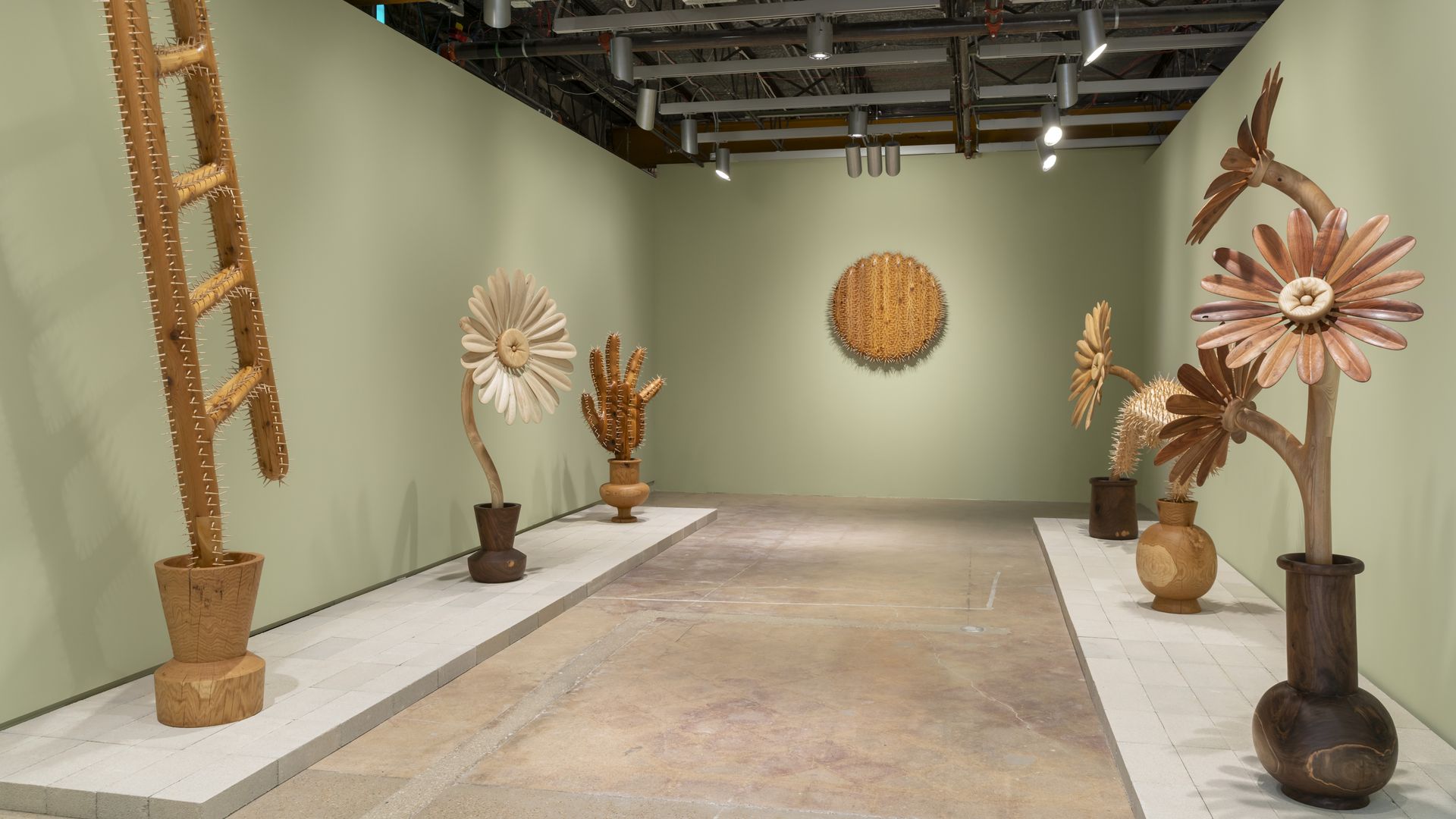 Gallery room with wooden sculptures resembling spiky plants and large flowers in various shades of brown and beige, displayed on platforms against light green walls.