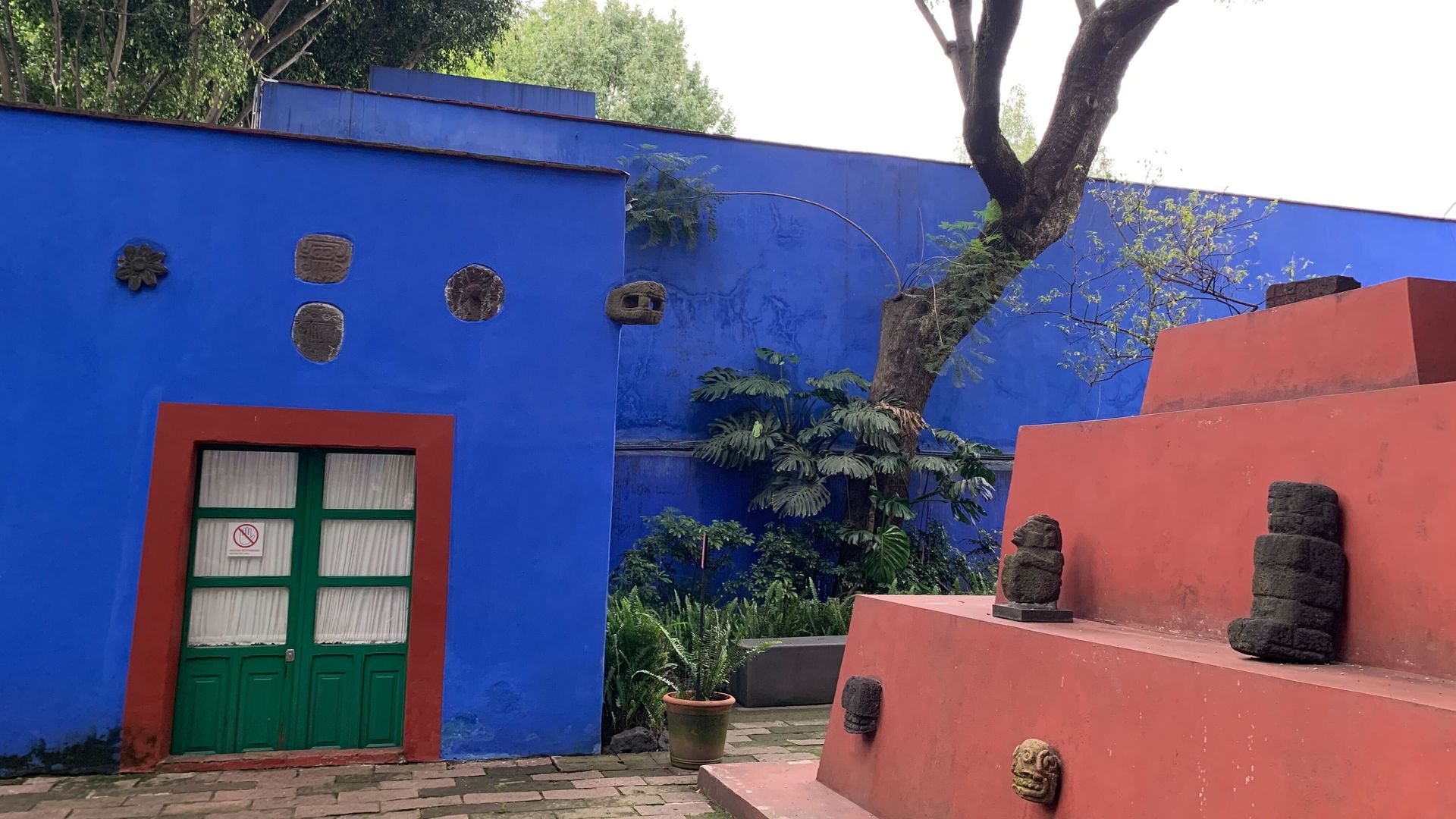 Outdoor scene with bright blue walls, a green door with a red frame, a red stepped monument adorned with stone sculptures, large tree, and green plants around the courtyard.