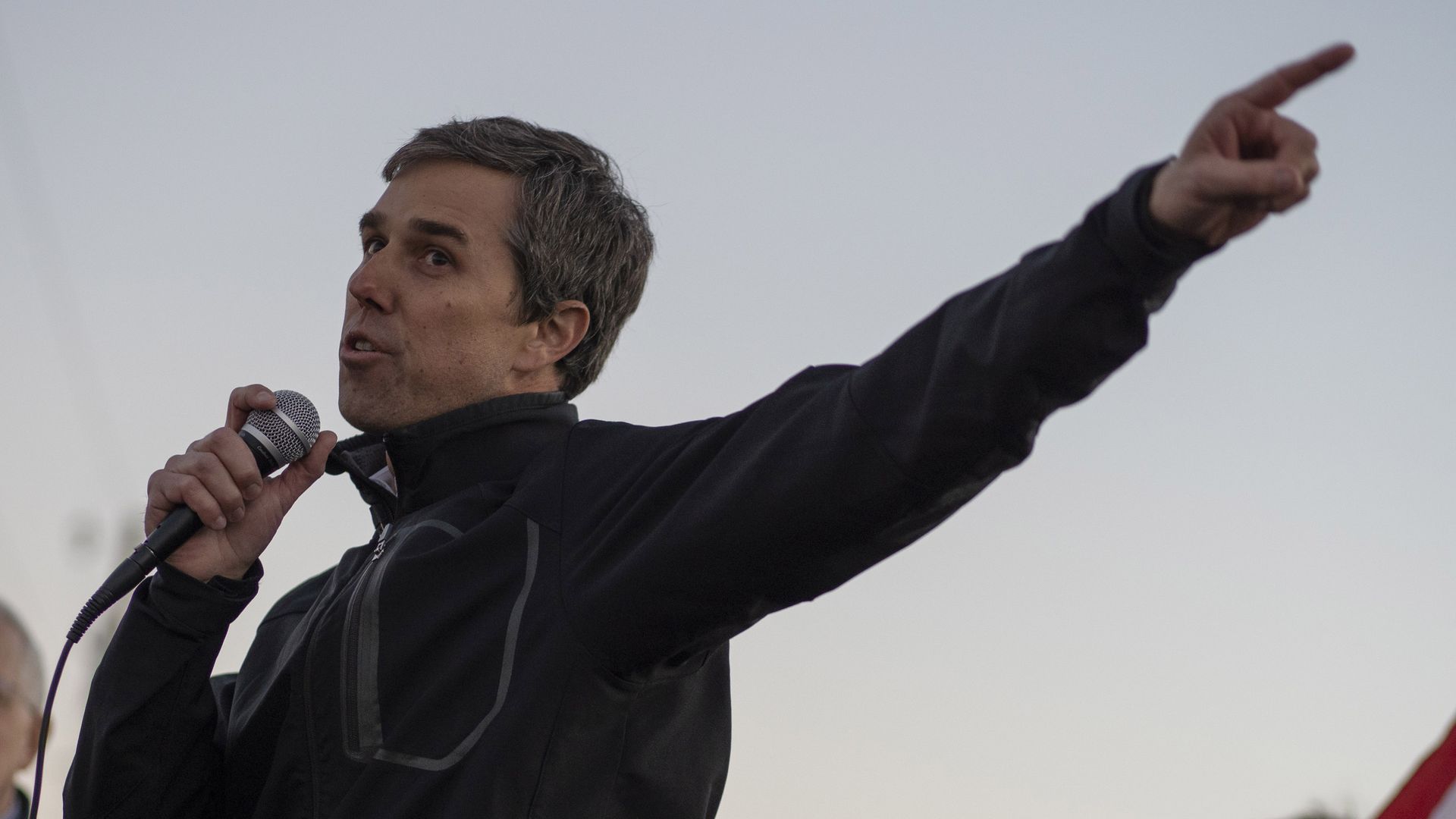 Beto O'rourke stands and speaks into a microphone at a rally, pointing to his left. 