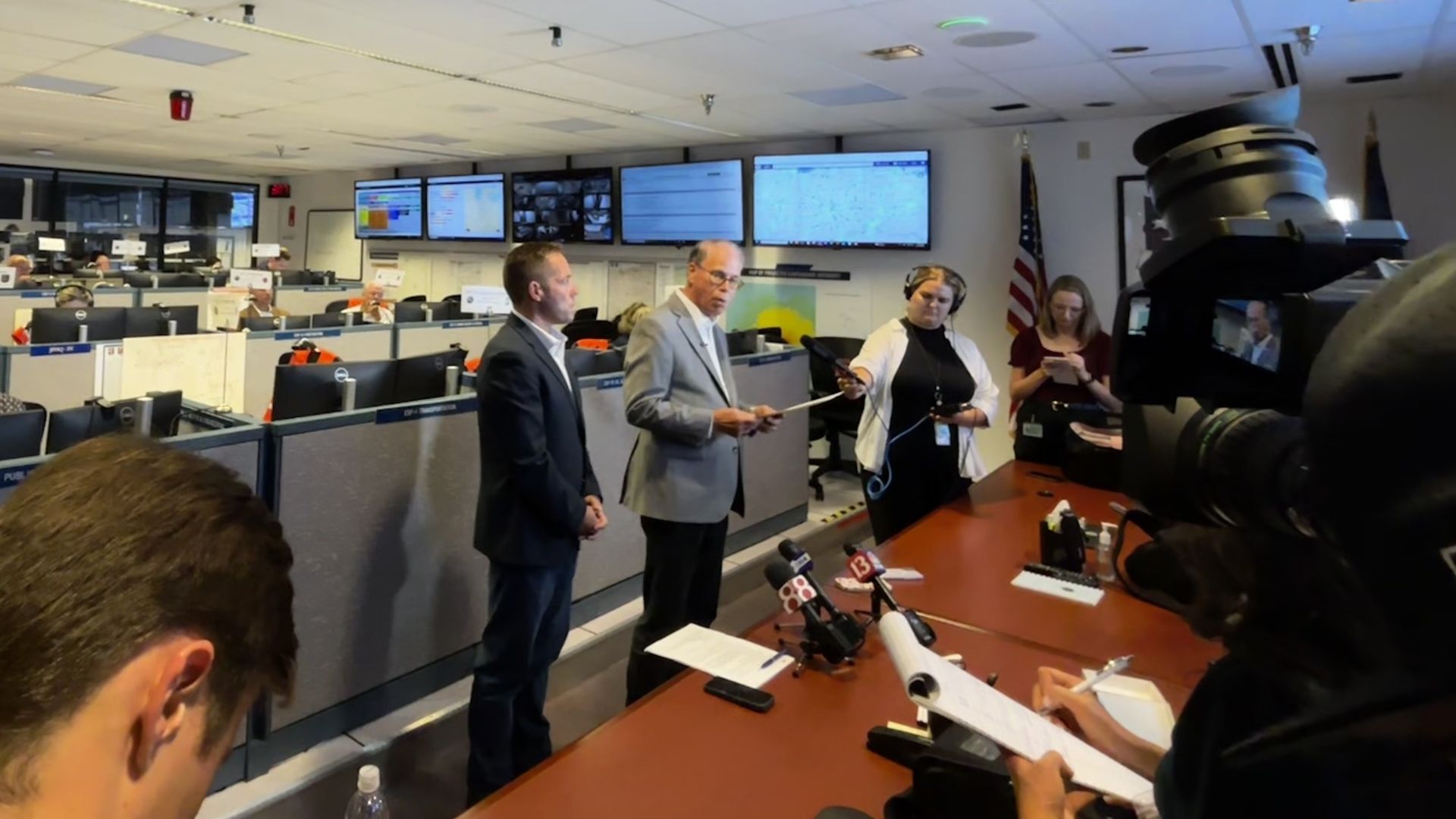 Press briefing in a busy operations room with row of desks and wall-mounted monitors. Three speakers in suits stand at a table with microphones; a large camera is in the foreground; American flag visible.
