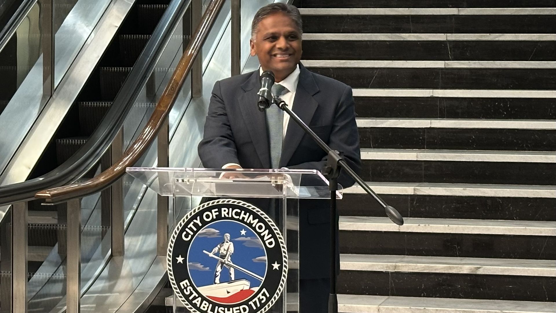 Smiling man in a dark suit speaks at a podium with two microphones, in front of the circular City of Richmond seal against black marble stairs; escalators are visible to the left.