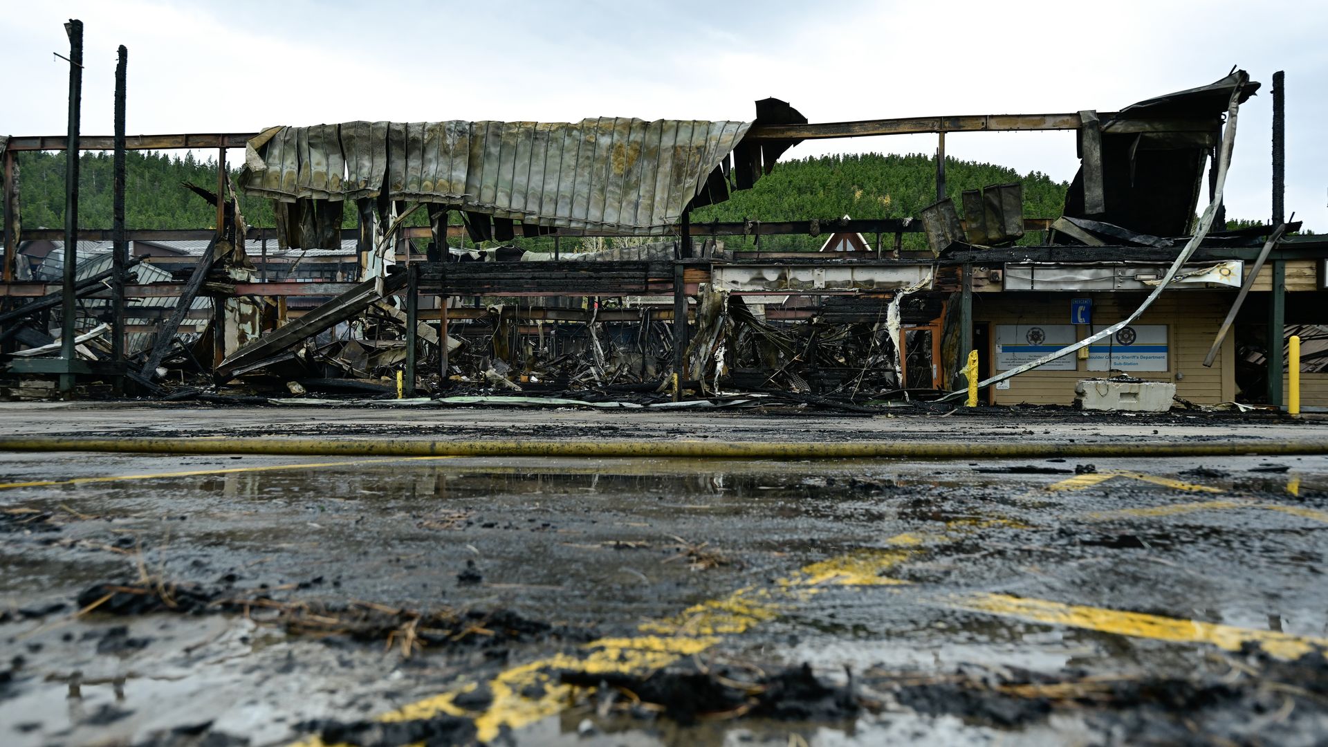 Burned and collapsed structure with charred beams and melted roof, surrounded by wet ground and yellow parking lines, with green forest and overcast sky in the background.