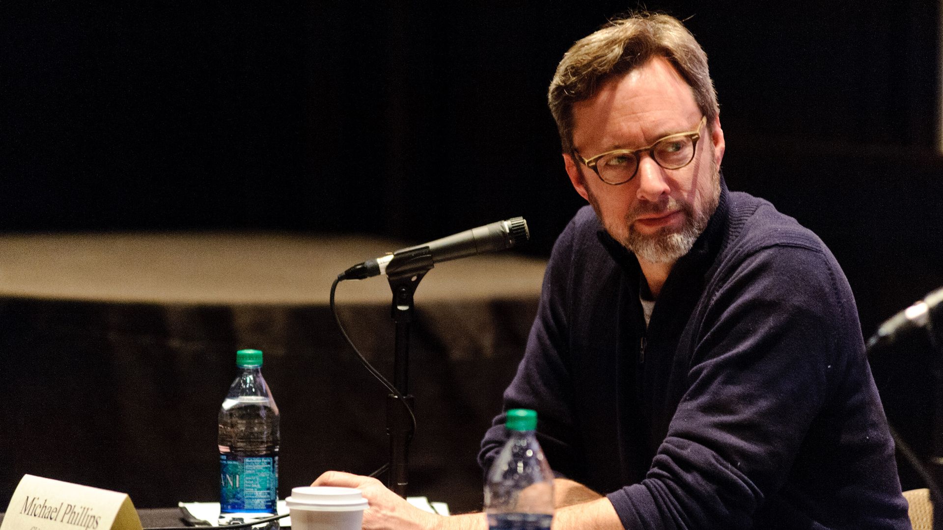Man with glasses and beard in navy sweater sitting at a panel table with microphone, water bottles, and a coffee cup, looking to the side during an event.